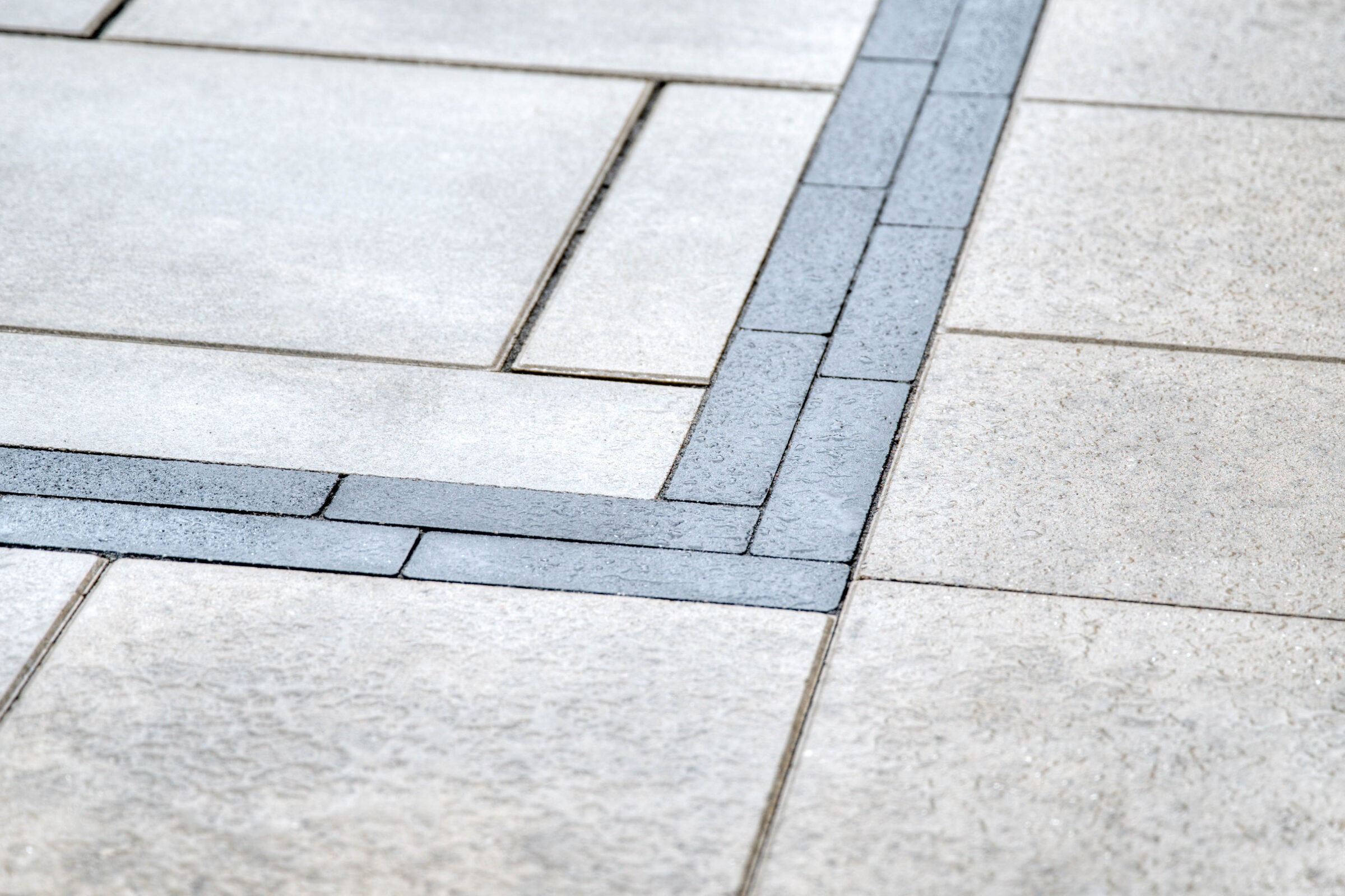 Close-up view of wet stone pavement, featuring light and dark gray tiles arranged in a geometric pattern. No people, landmarks, or buildings visible.