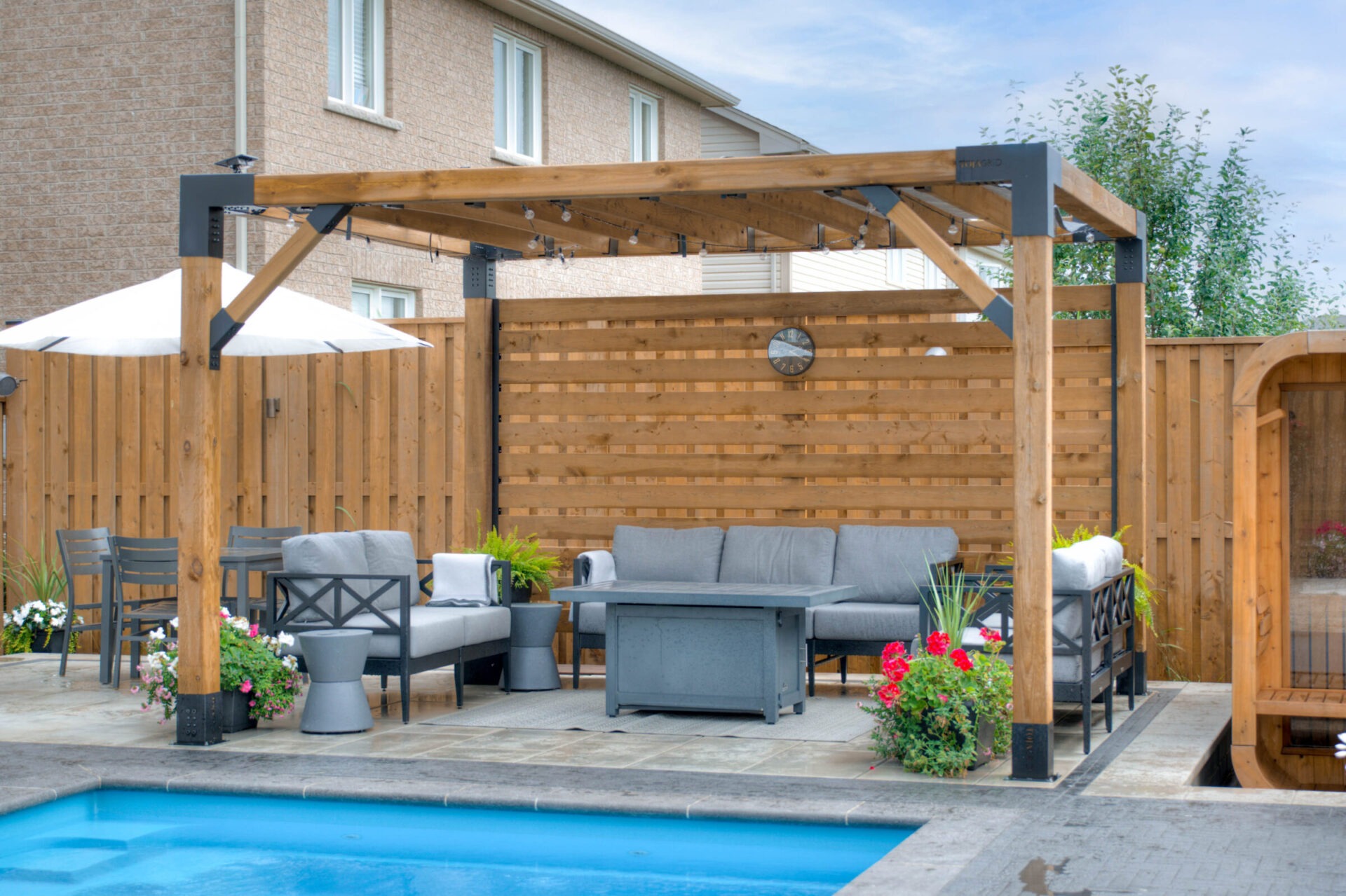A backyard features a cozy seating area under a wooden pergola, next to a pool. Bright flowers add color to the scene.