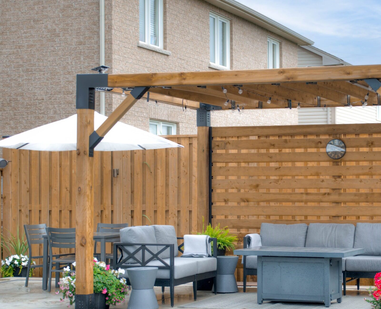 Poolside patio with a wooden pergola, gray seating, and decorative plants. Enclosed by a wooden fence, brick house in background.