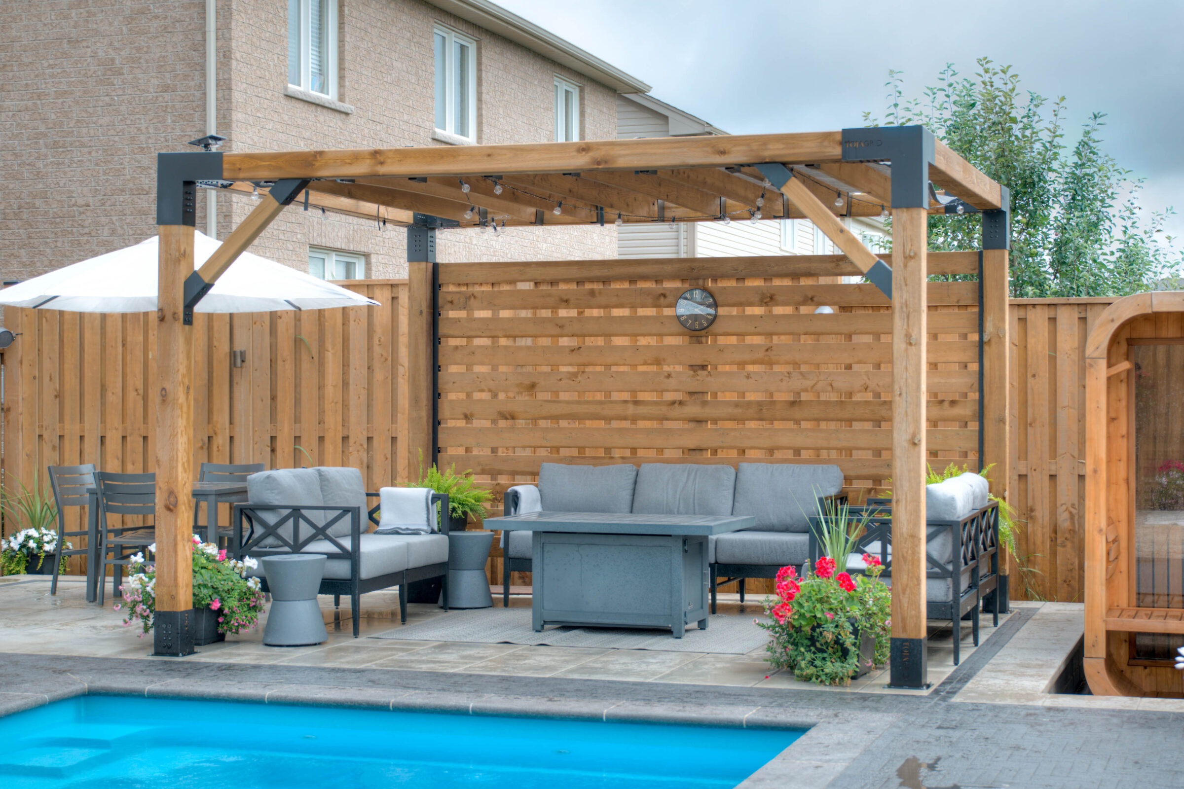 A backyard features a wooden pergola, gray seating area, and blue pool, all surrounded by a privacy fence and lush greenery.