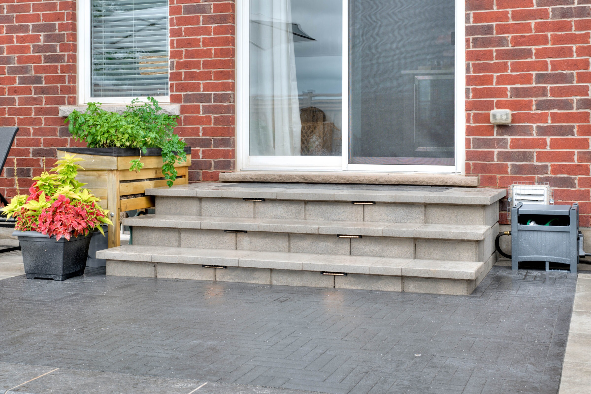 Brick house with sliding glass door opening onto stone steps. Planters with vibrant foliage adorn the patio area, featuring slate paving.