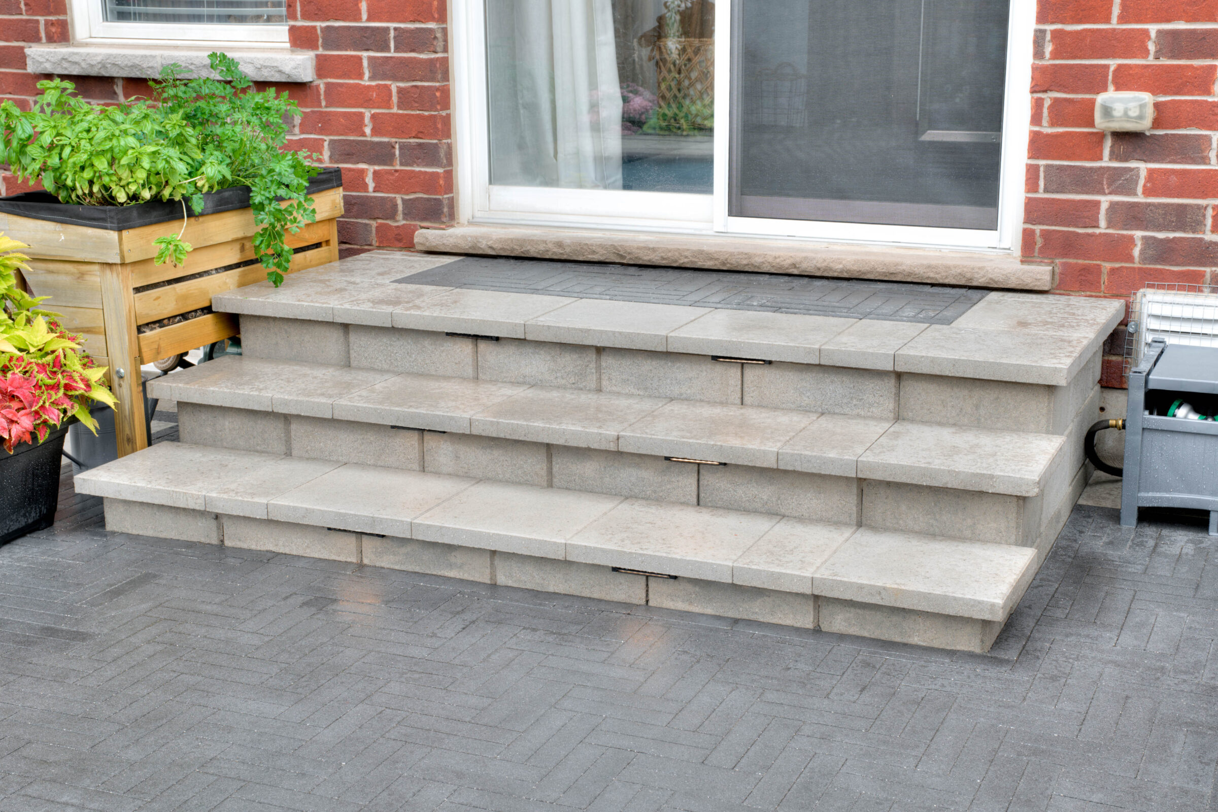 Concrete steps lead to a brick house's sliding glass door, flanked by potted plants and a wooden planter box.