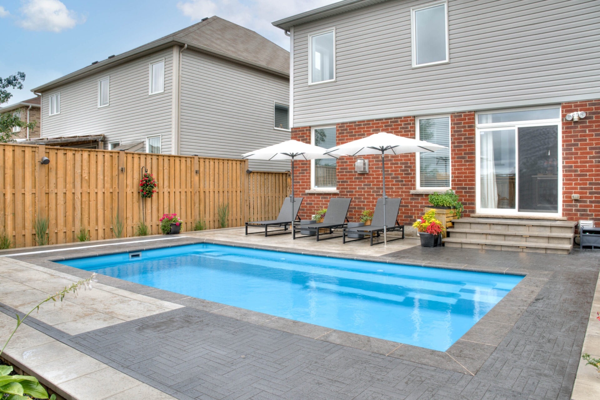 Backyard view with a rectangular swimming pool, surrounded by patio chairs and umbrellas, against a wooden fence and modern brick house exterior.