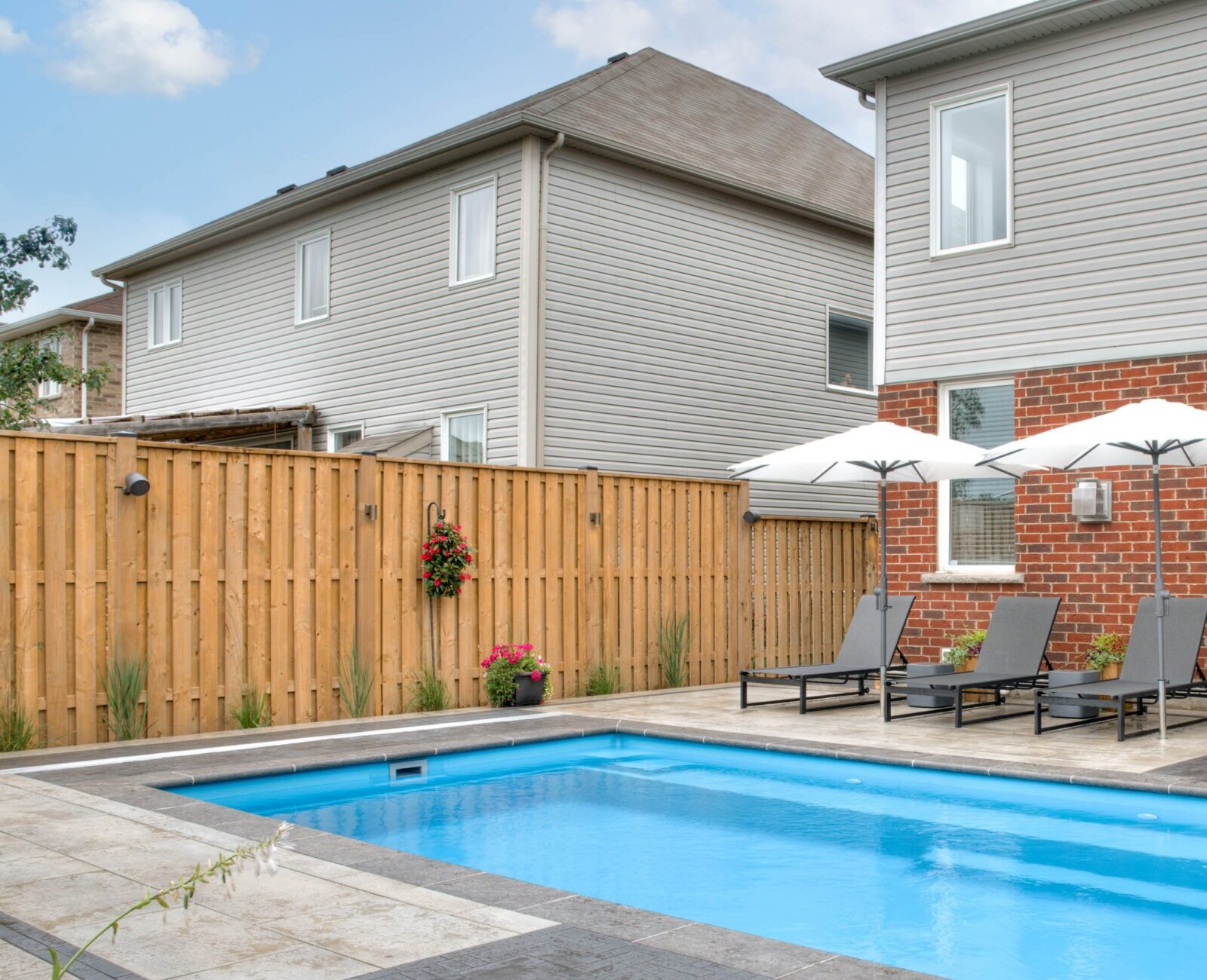 Modern backyard featuring a rectangular pool, surrounded by stone tiles, sun loungers under umbrellas, and a red brick house with large windows.