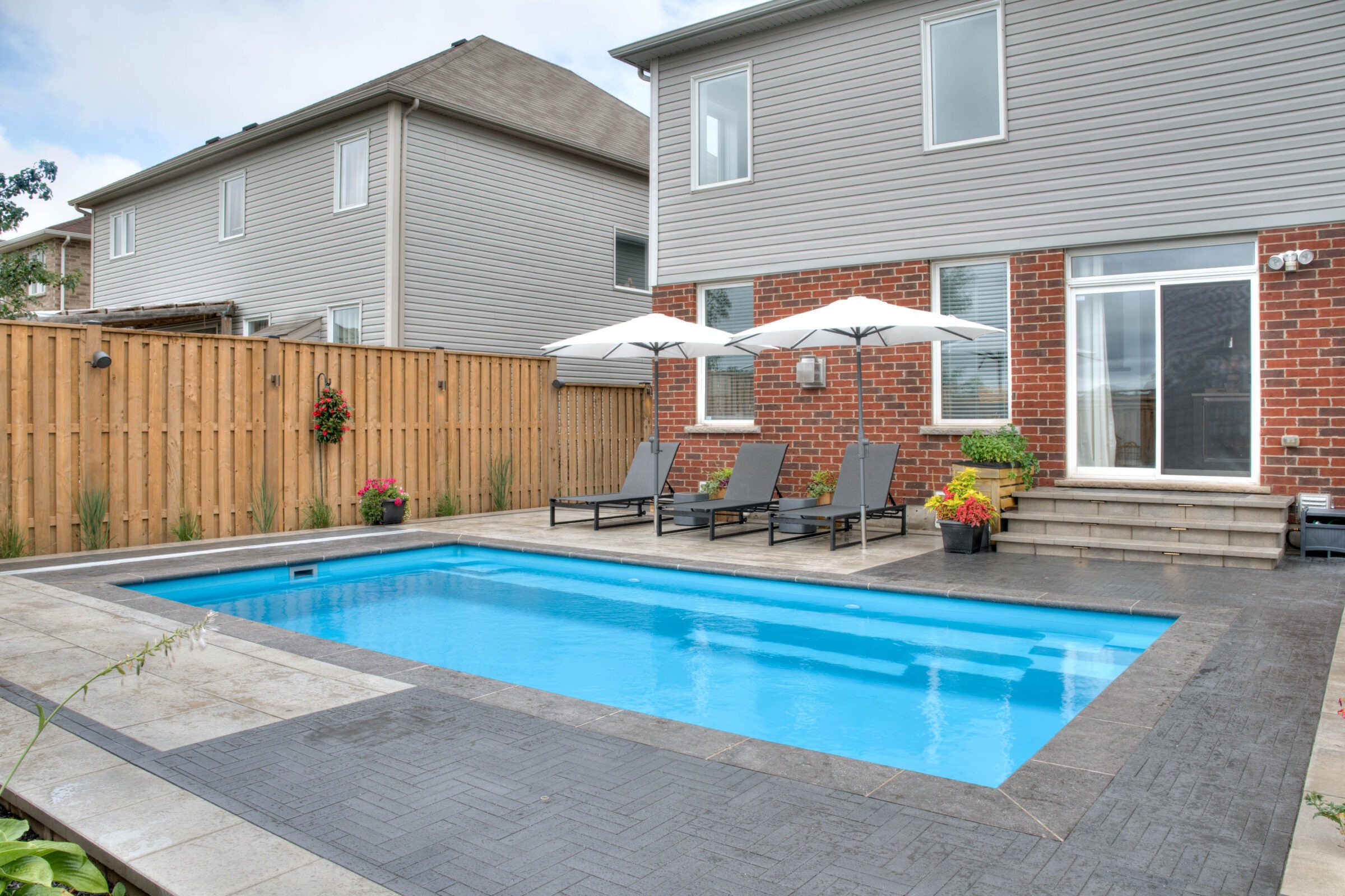 A backyard scene with a rectangular pool, brick house, wooden fence, gray lounge chairs, and white umbrellas. No people are present.