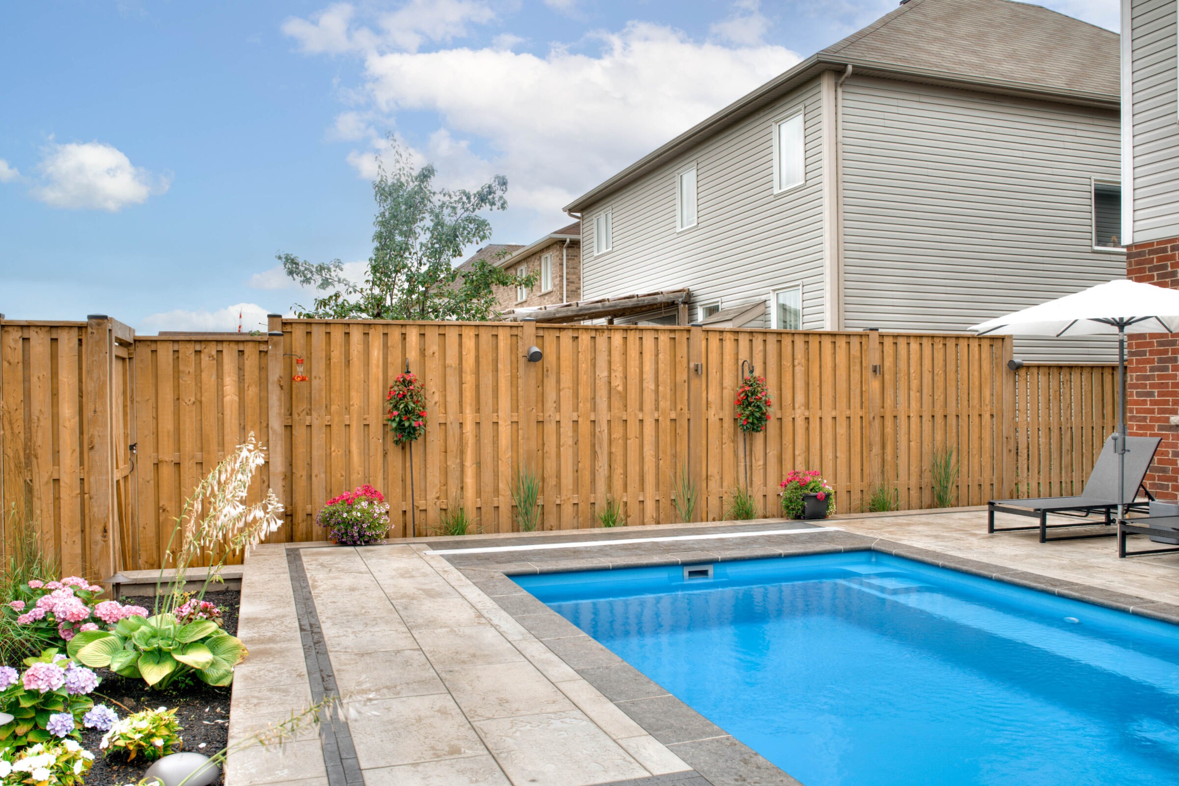 Backyard with a swimming pool, lounge chairs, umbrella, wooden fence, and colorful flowers. Houses in the background under a partly cloudy sky.