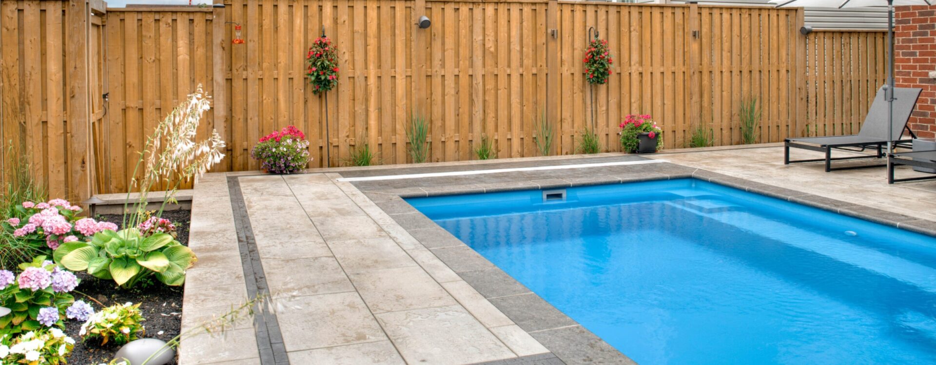A backyard features a rectangular blue pool, stone patio, wooden fence, lounge chairs, and vibrant flowers in planters, creating a serene atmosphere.