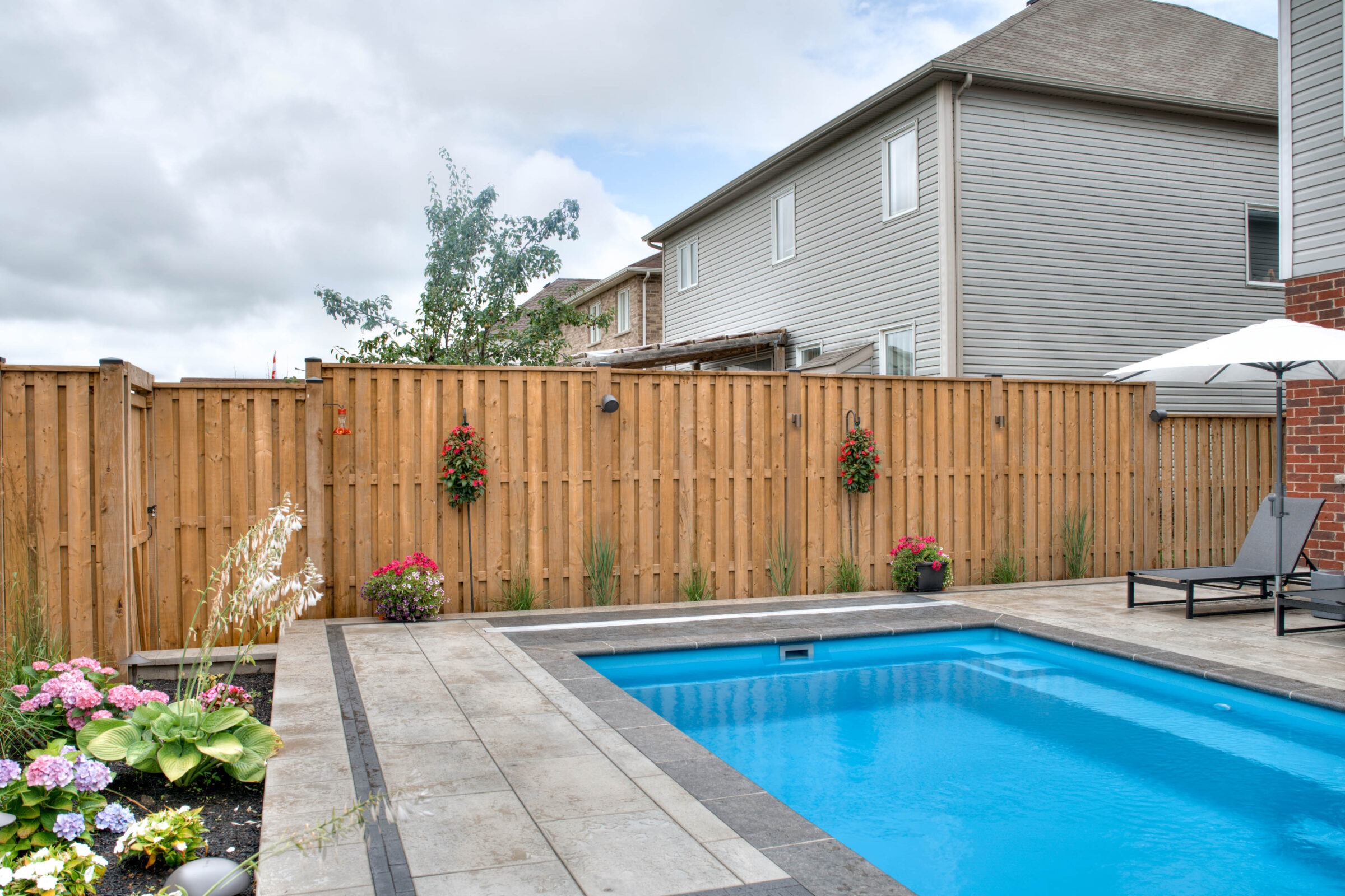 A backyard with a swimming pool, lounge chairs, umbrella, wooden fence, and vibrant flowers under a cloudy sky near residential houses.