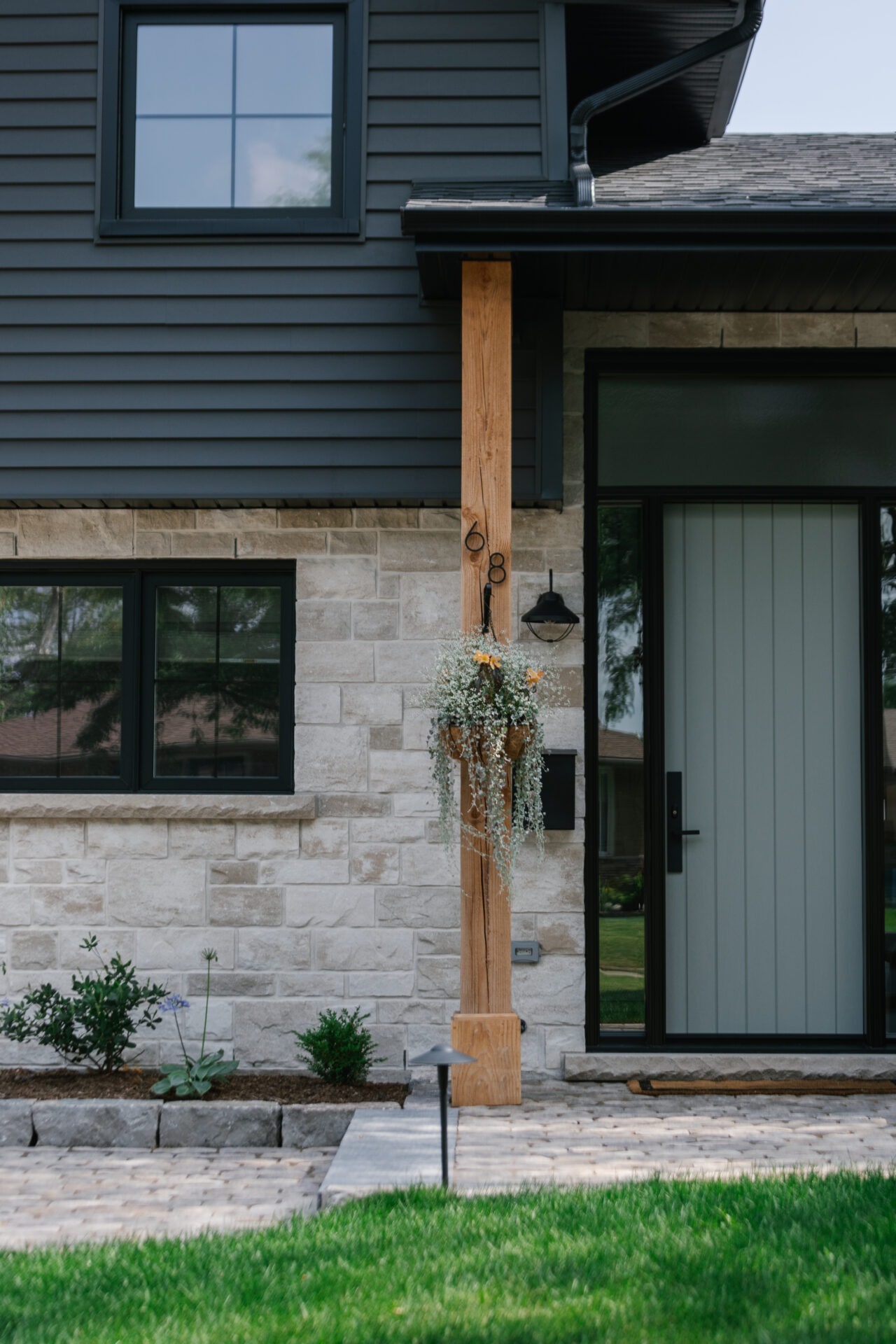 Modern house exterior with stone and siding, featuring a wooden post with number 68 and hanging plants, alongside a neatly landscaped walkway.