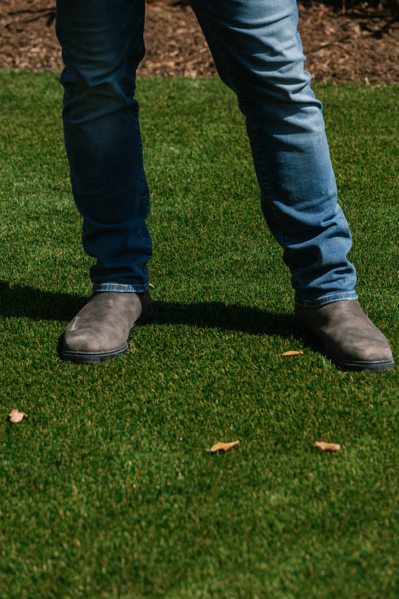 A person is standing on green grass, wearing blue jeans and brown boots, with scattered dry leaves around.