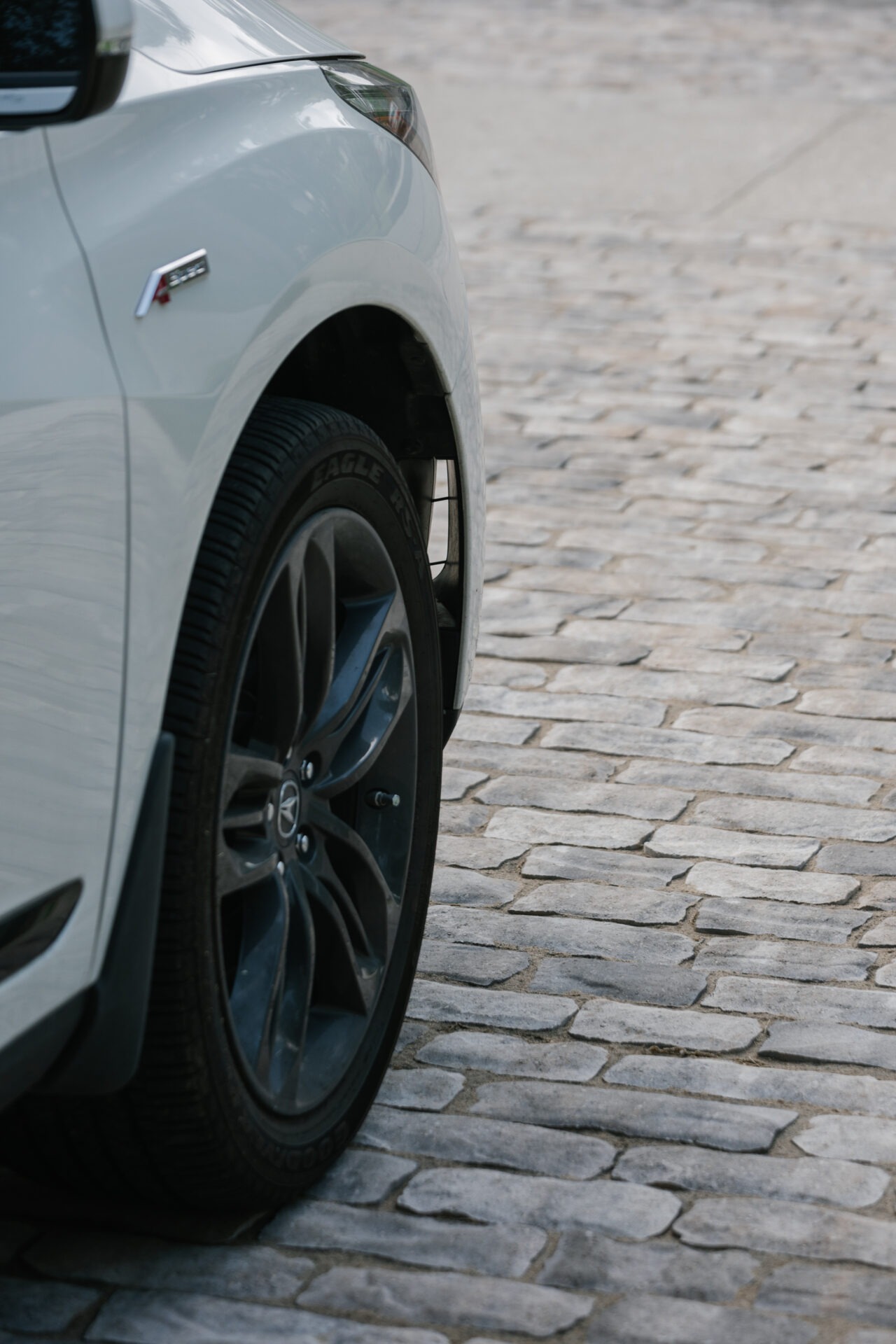 A white car parked on cobblestone street, showcasing the front wheel and part of the fender with no recognizable landmarks visible.