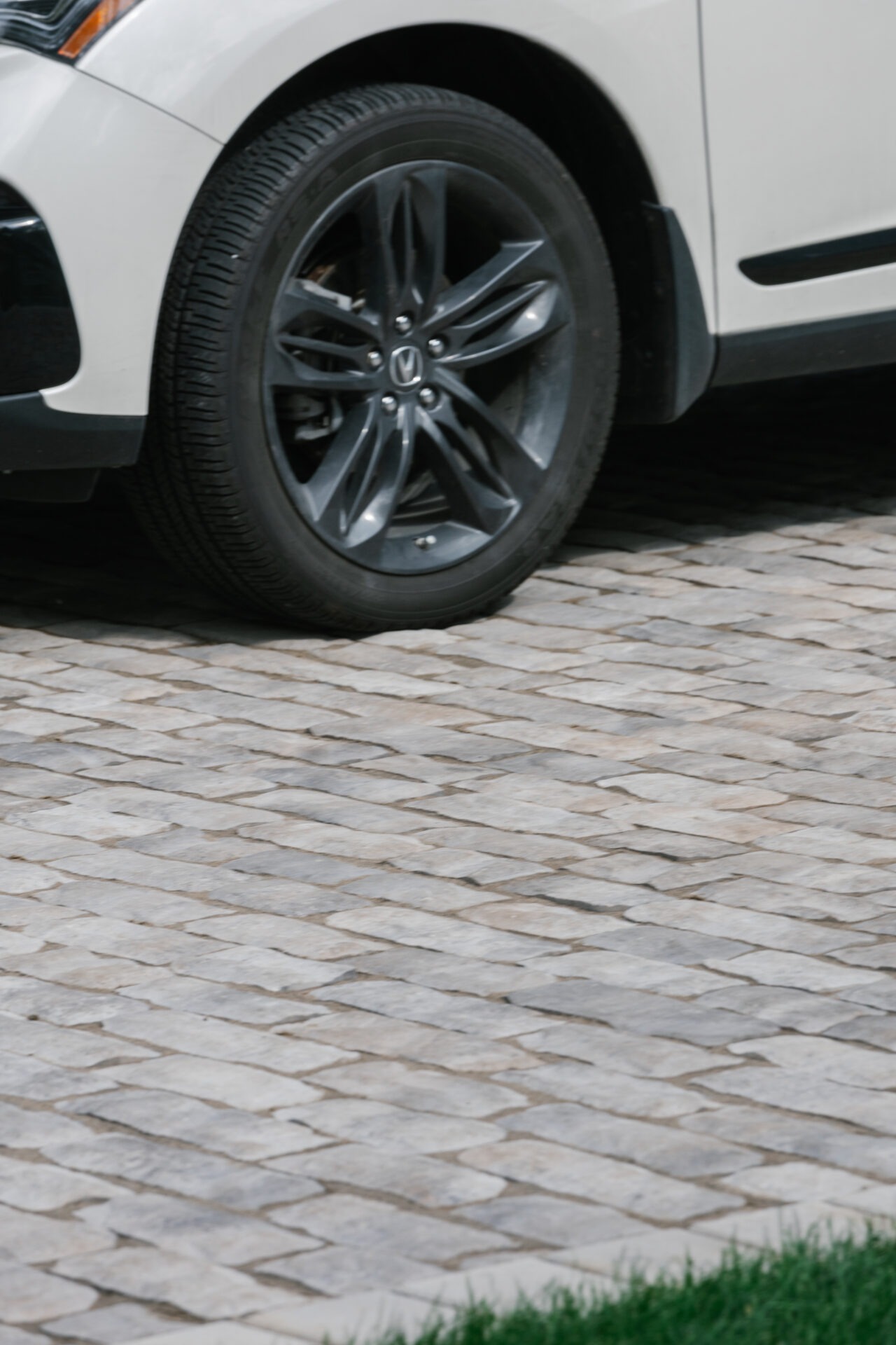 A white car is parked on a cobblestone driveway, showing its front wheel and a grassy edge in the foreground.