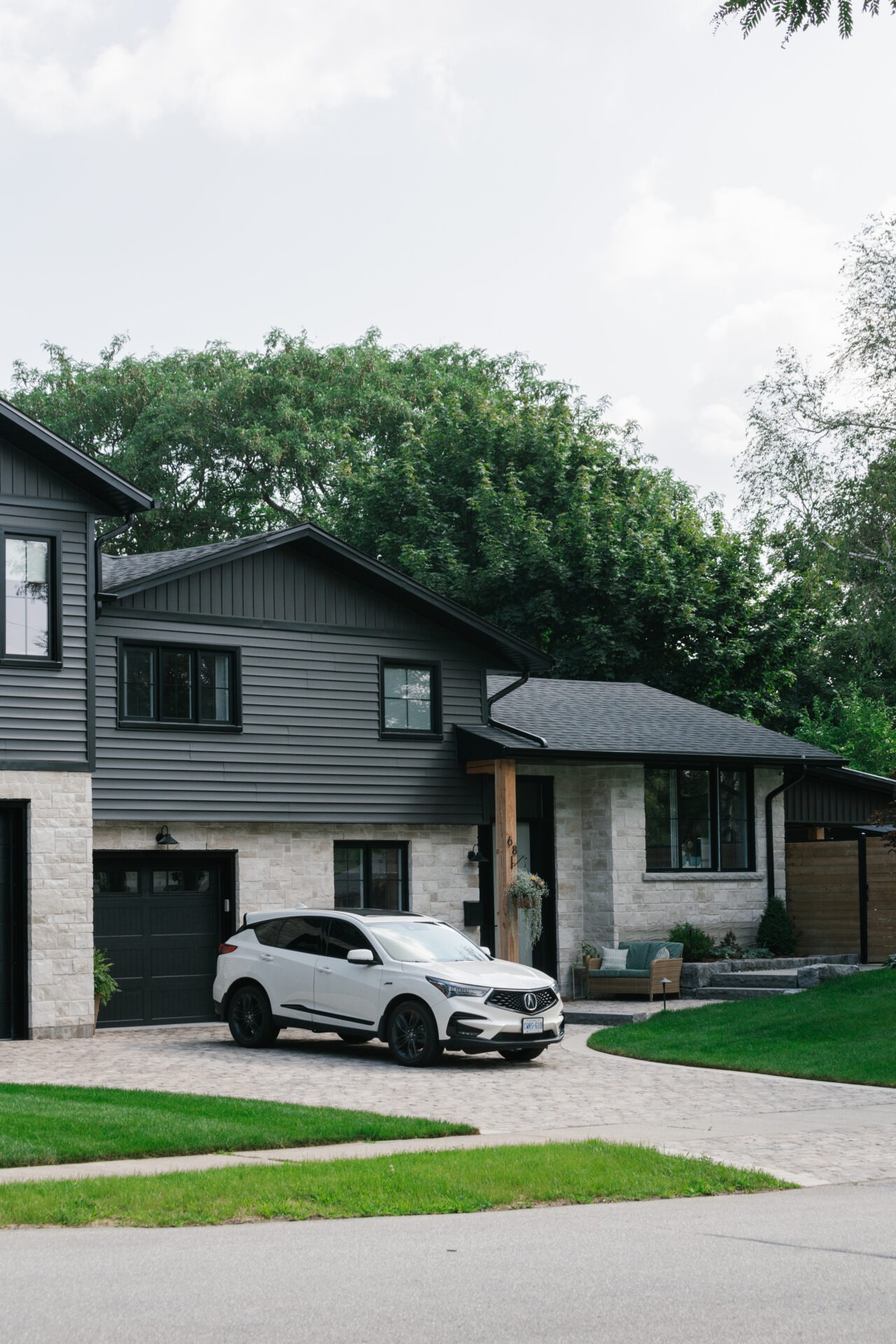 Modern suburban house with dark siding and stone exterior, featuring a parked white SUV on the driveway surrounded by lush green trees.