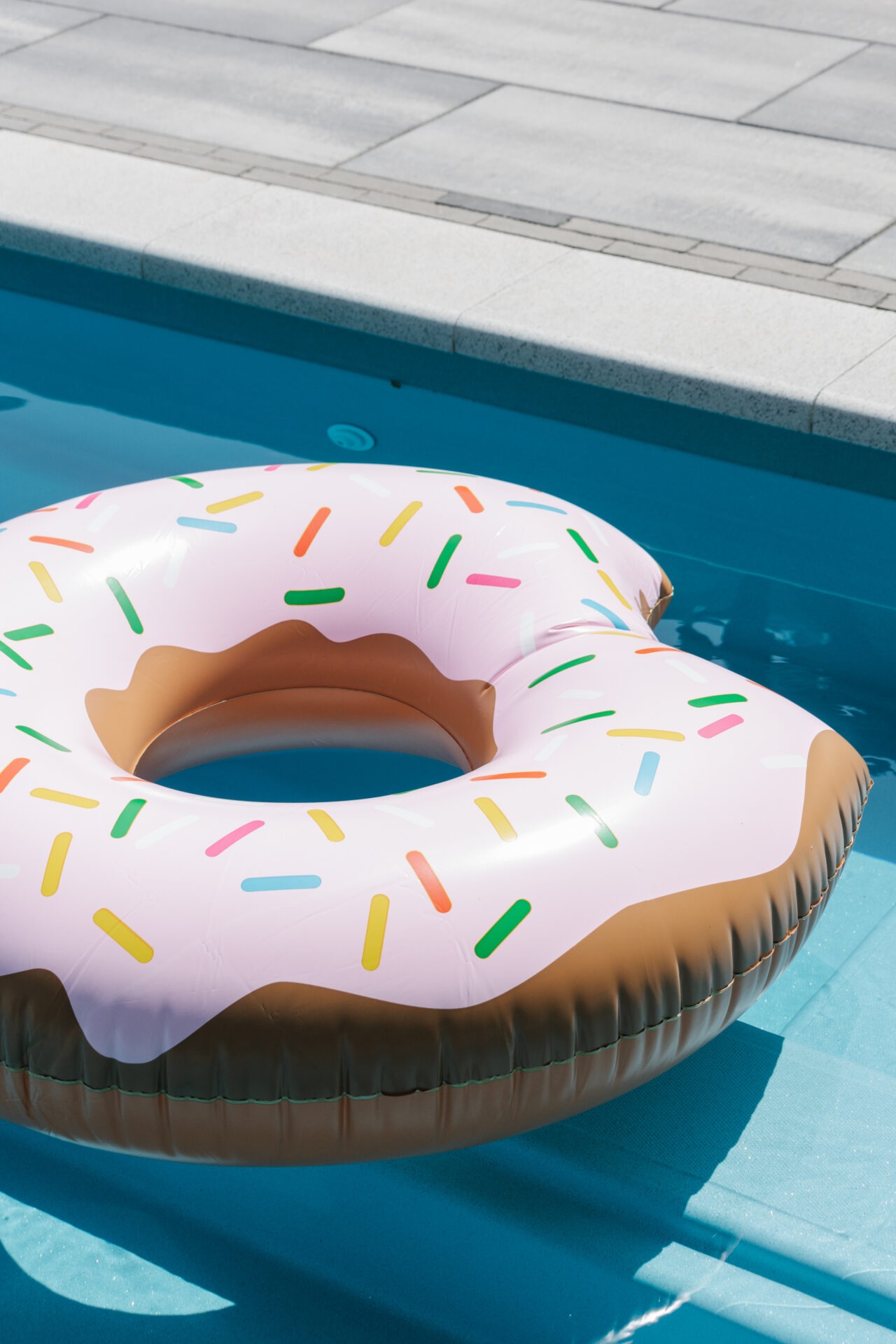Inflatable donut-shaped pool float, decorated with colorful sprinkles, floats on clear blue water beside a paved pool deck.