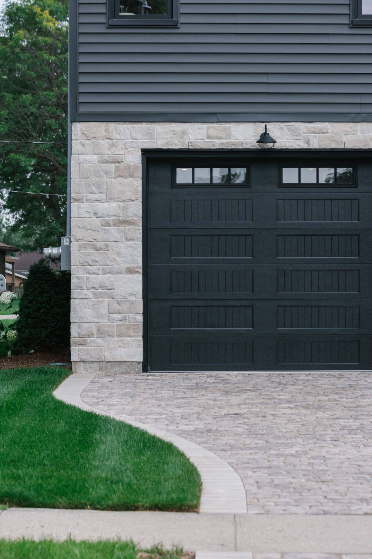 Modern garage with dark door, stone wall, and paving leading to neat, green lawn. Simple exterior, minimal decoration, suburban setting.