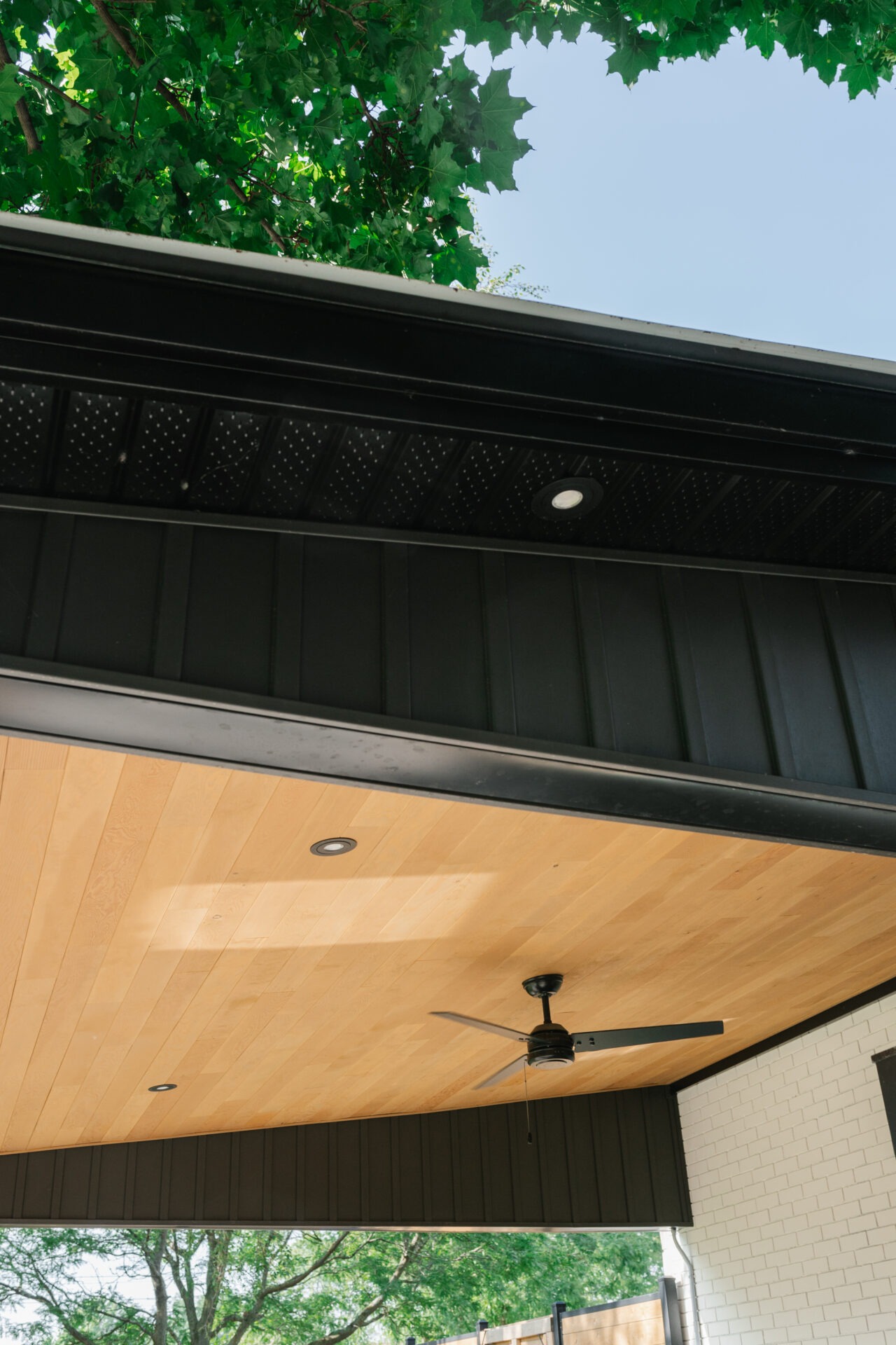 Modern ceiling with a wooden finish, recessed lighting, and a ceiling fan, beneath green leafy tree branches against a clear sky.
