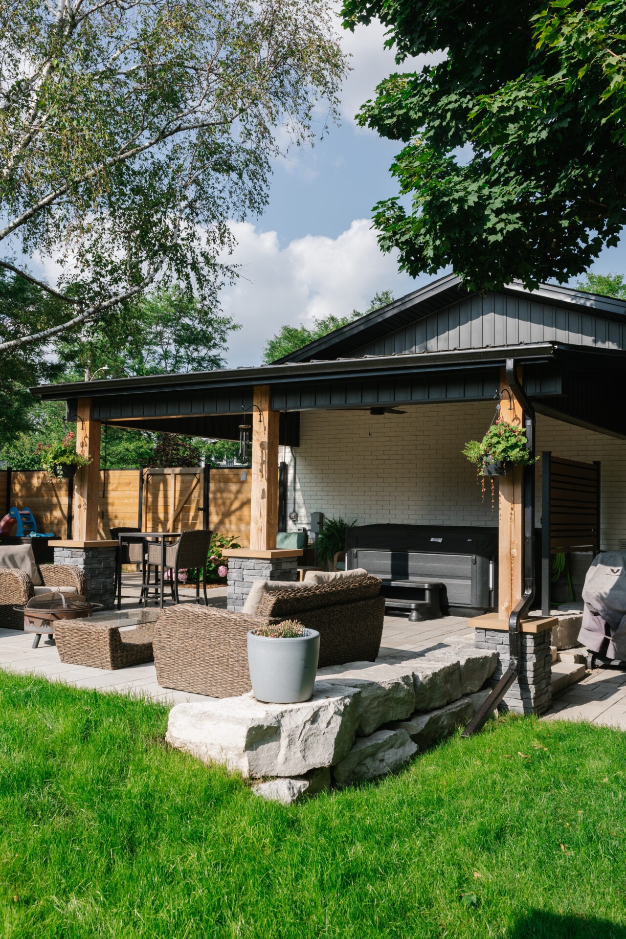 A cozy backyard patio with wicker furniture, lush green lawn, a modern pergola, and hanging plants under a clear blue sky.