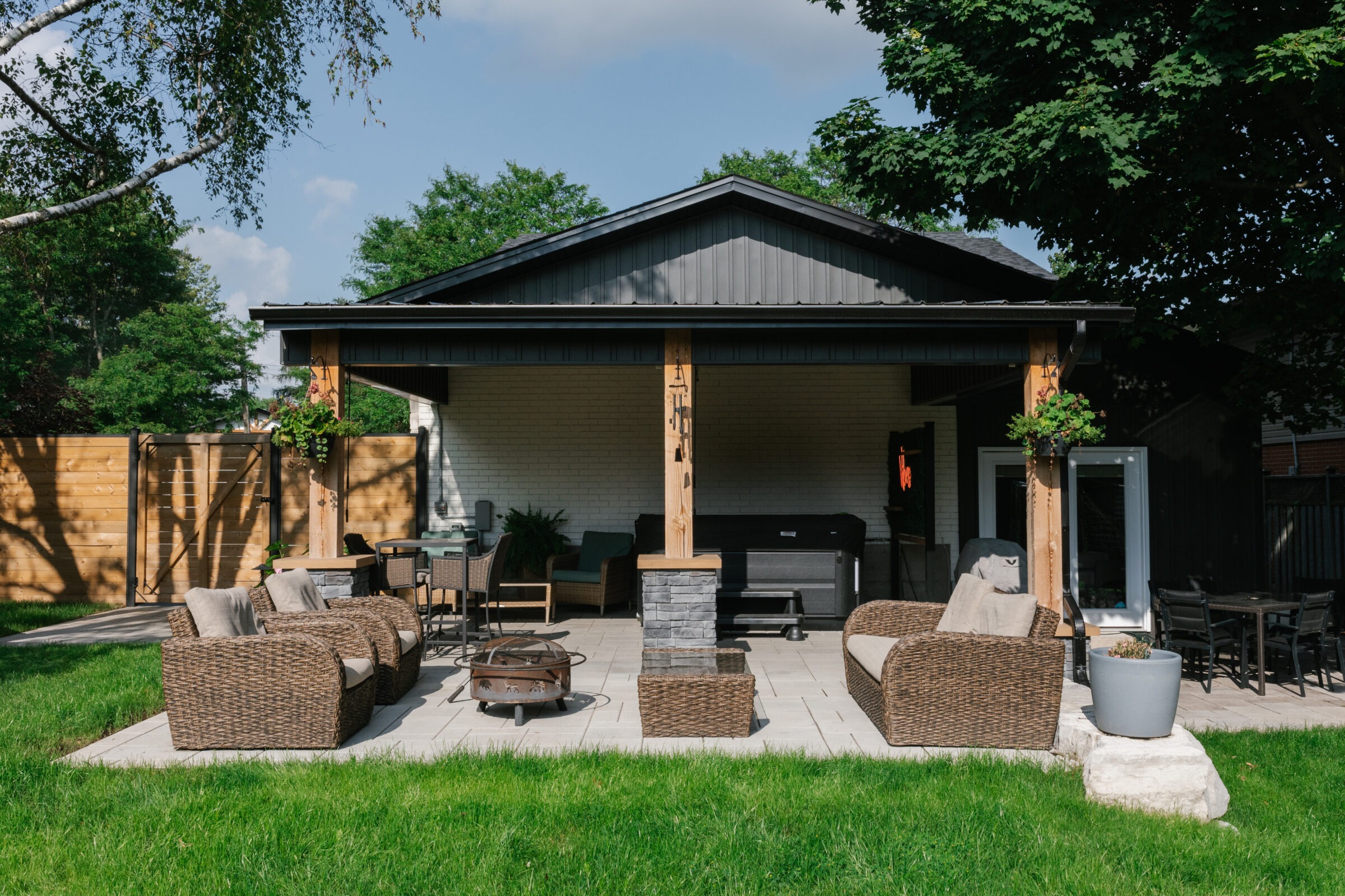 A cozy patio with wicker furniture, fire pit, and garden view. Wooden beams support a roofed structure, surrounded by lush green trees.