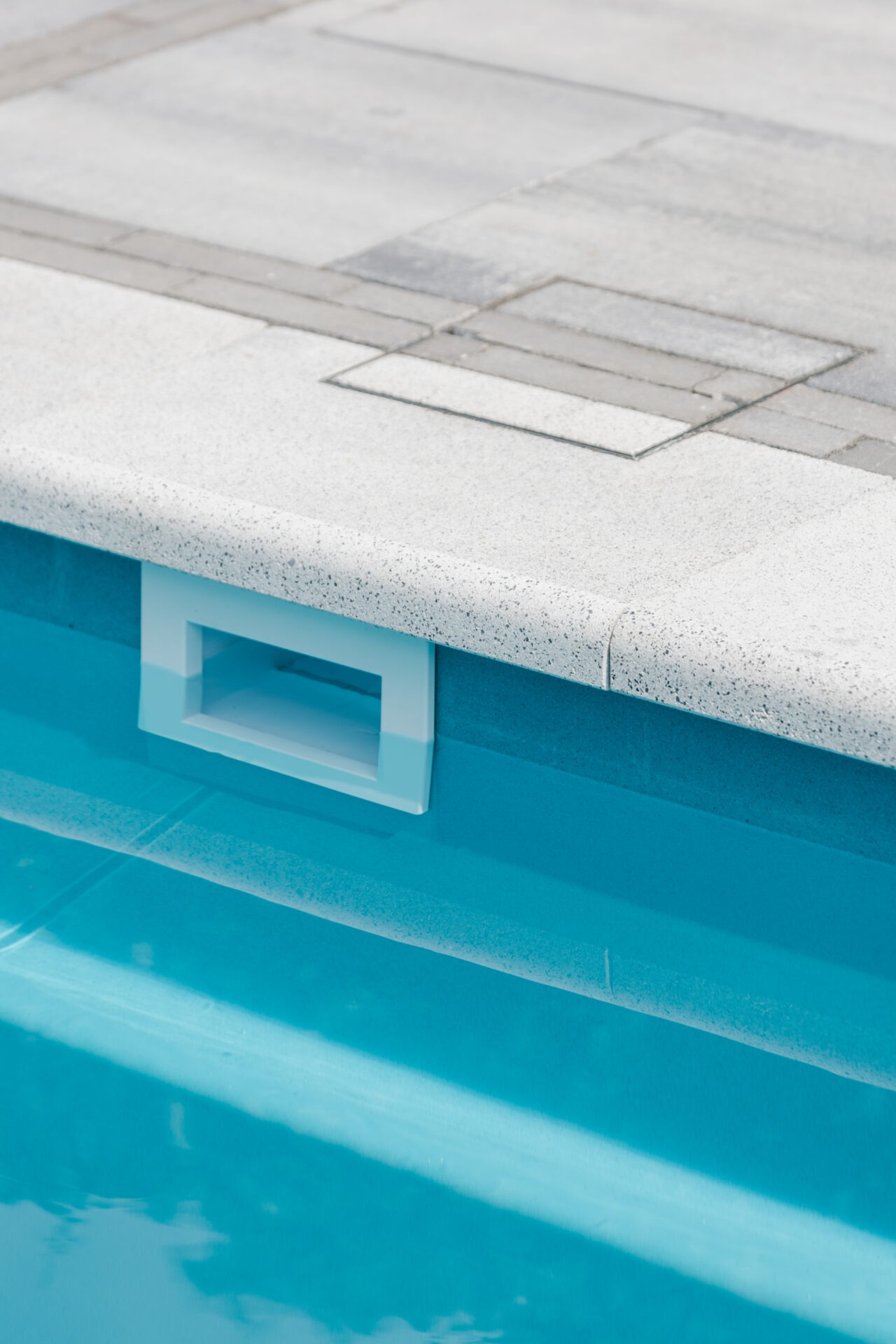 Close-up of a pool skimmer in a swimming pool with light blue water and a stone-tiled pool edge.