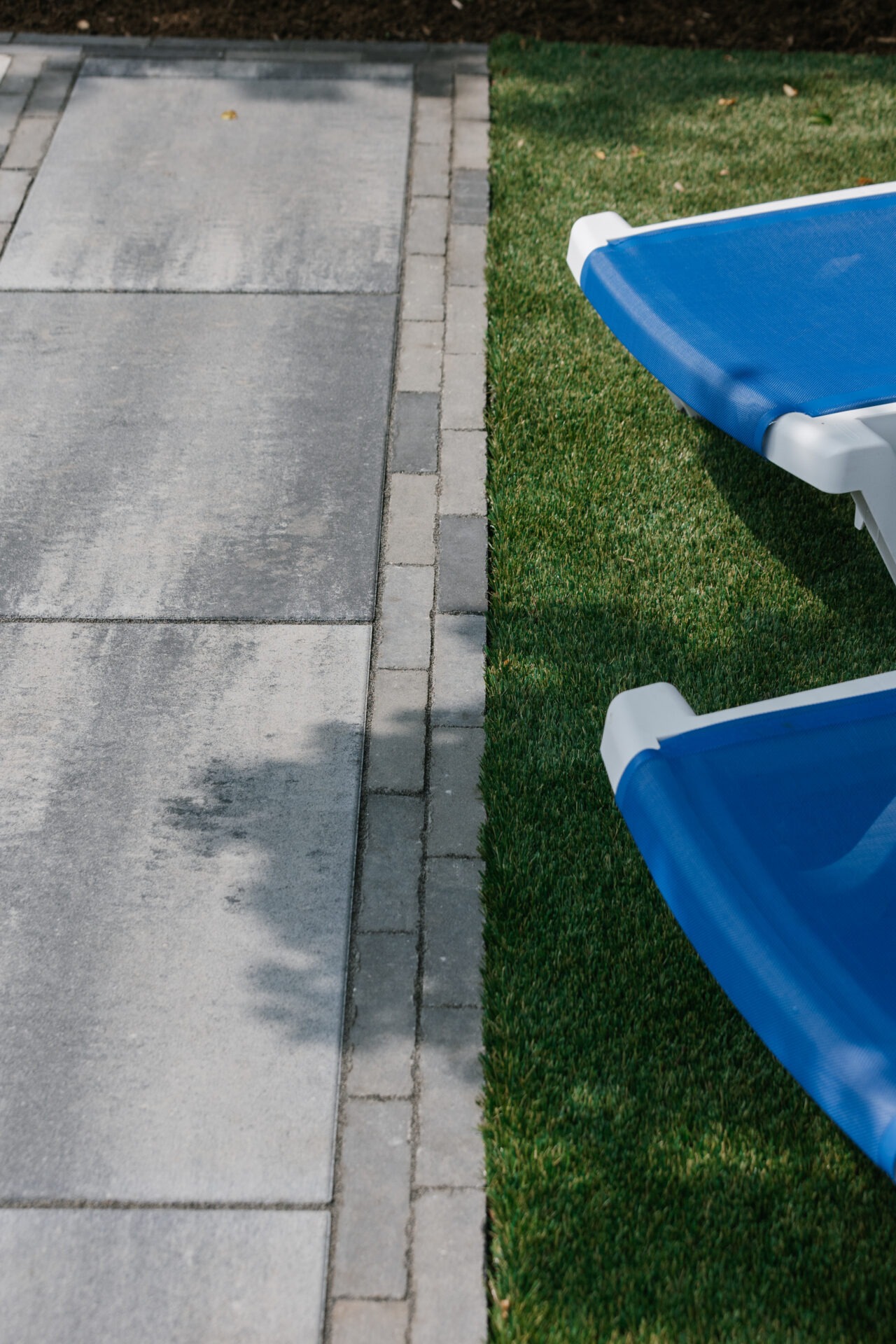 A blue lounge chair on green grass beside a gray stone pavement, casting a shadow in bright sunlight.