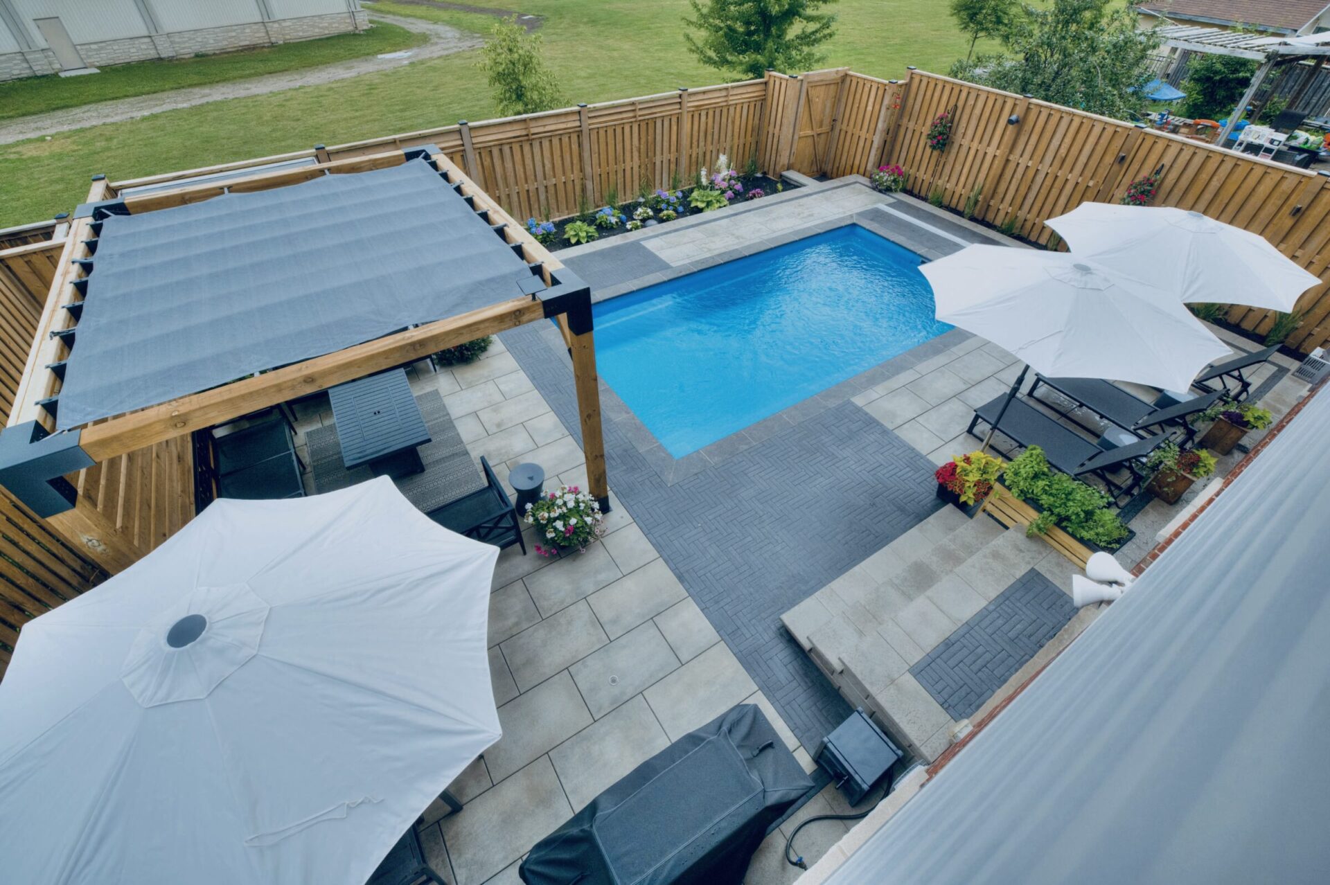 A backyard with a blue pool, patio furniture, pergola, and white umbrellas, surrounded by a wooden fence and landscaped garden.
