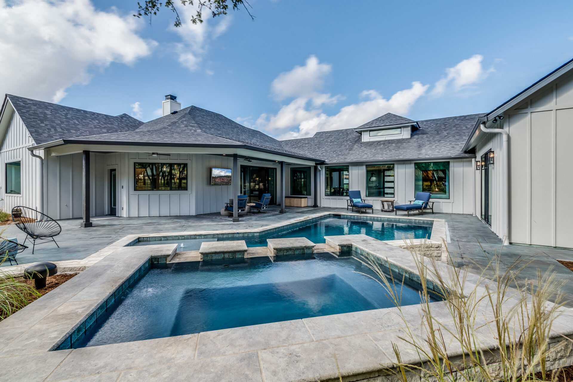 Modern house with gray roof and pool in open courtyard, featuring blue lounge chairs and glass doors under a clear, blue sky.