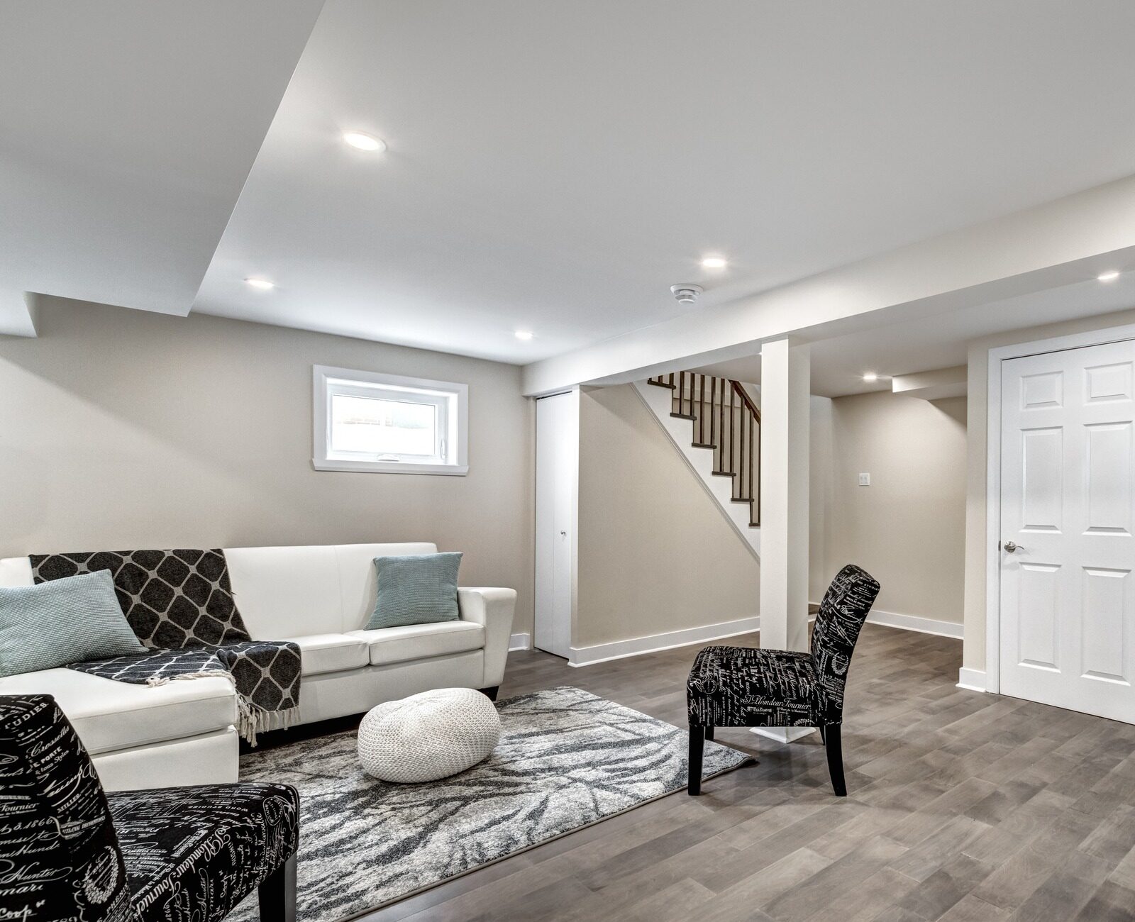 Modern basement with white sofa, patterned chairs, and a grey rug. Wooden stairs lead upward. Neutral colors create a cozy atmosphere.
