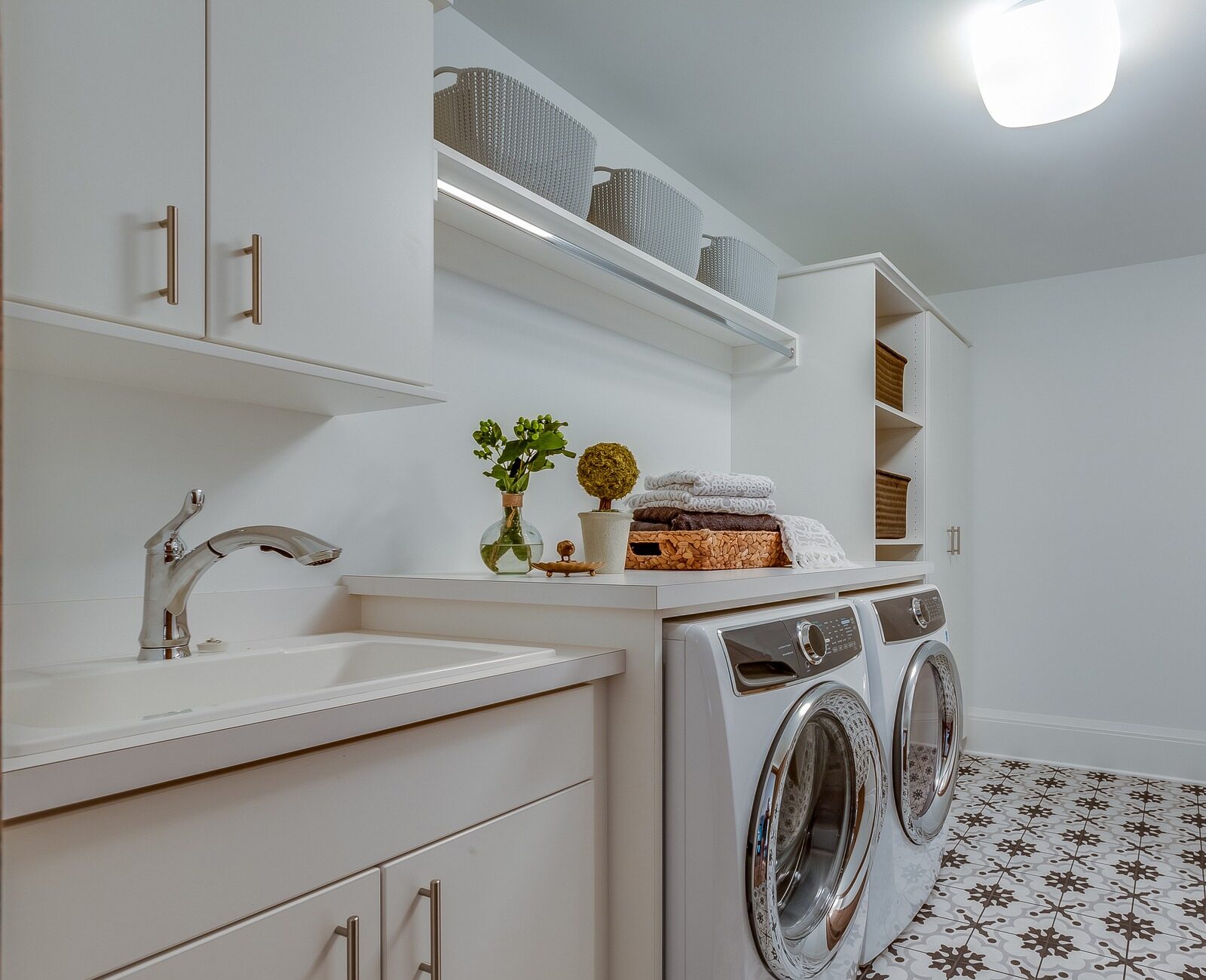 A tidy laundry room with patterned floor tiles, a washer, dryer, sink, and storage shelves, featuring baskets and neatly folded towels.