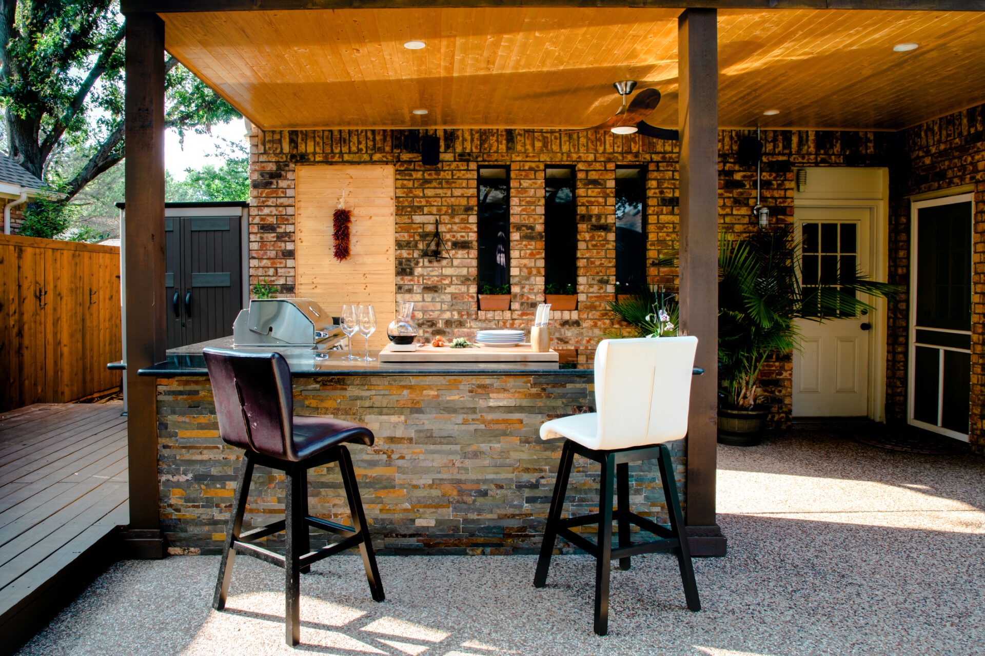 Outdoor kitchen with brick walls features a grill, bar counter, and two chairs. Plants adorn the area, creating a cozy atmosphere.