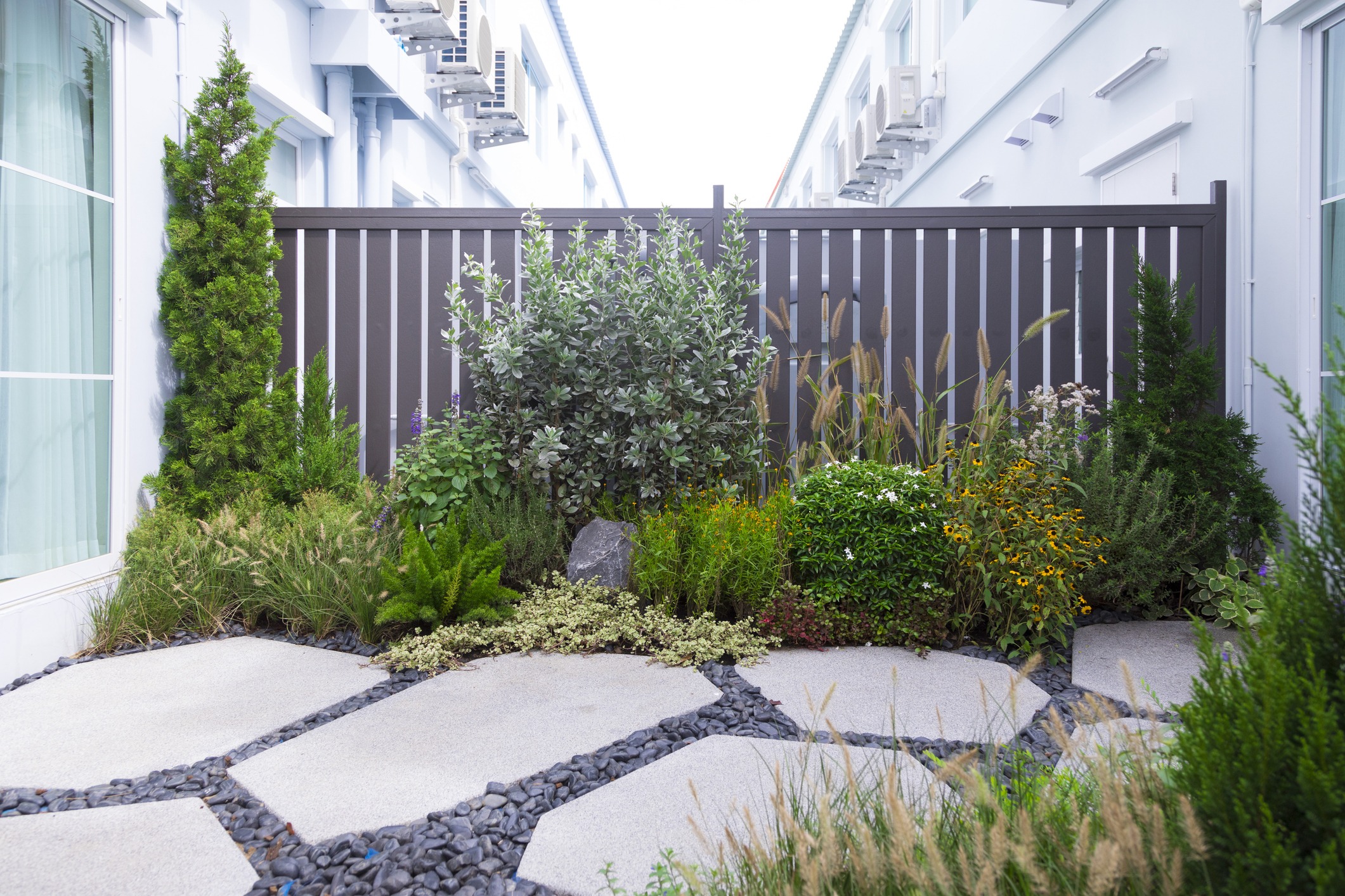 A small garden features diverse plants and shrubs, bordered by a modern fence, with white buildings and air conditioning units visible above.