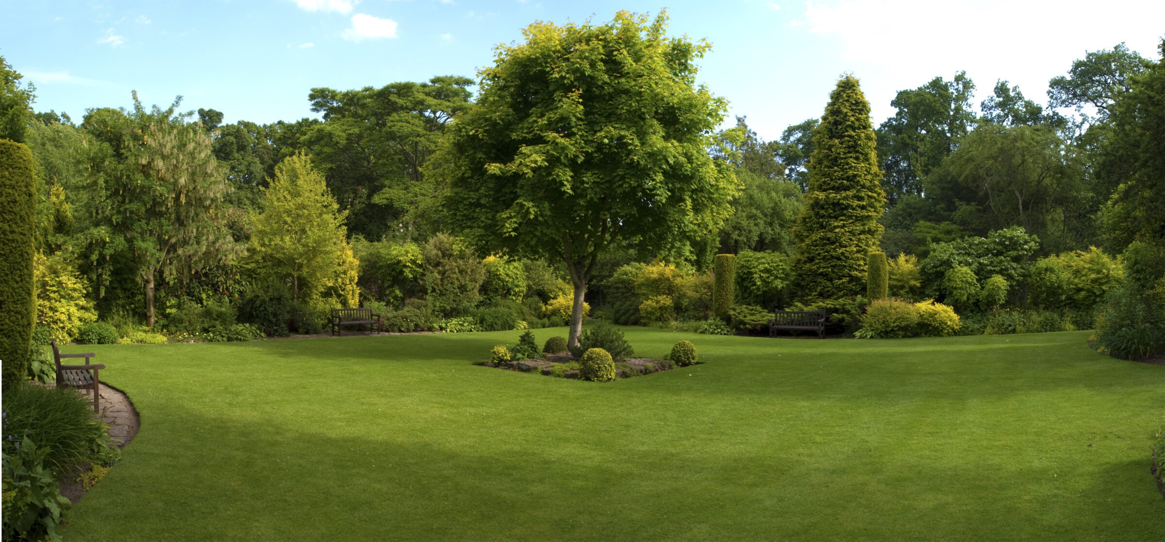 A well-maintained garden with lush green lawns, diverse trees, and shrubs, surrounded by benches, creating a peaceful natural setting under a clear sky.