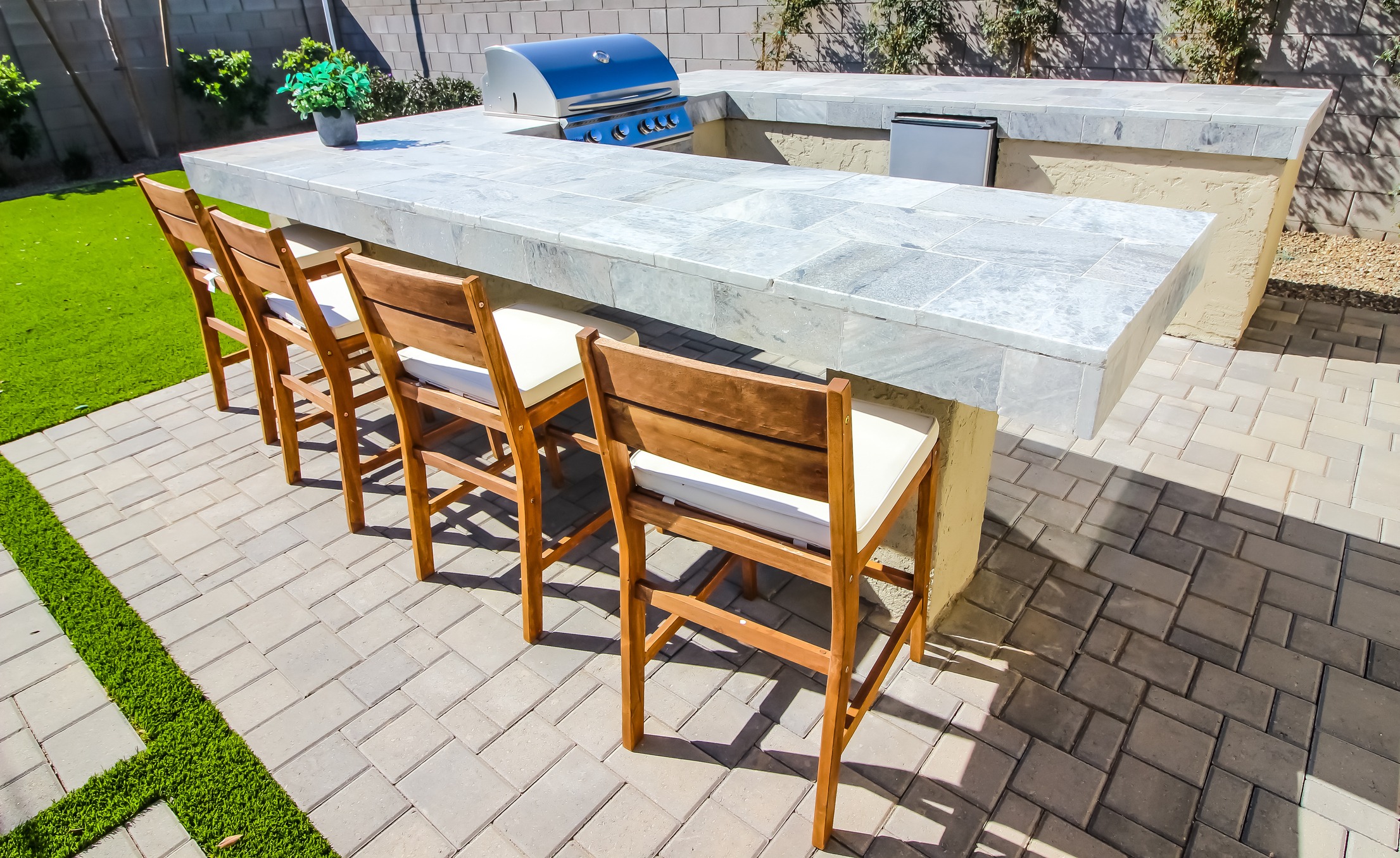 Outdoor patio kitchen with stone countertops, four wooden barstools, and a blue barbecue grill. Paved flooring and green artificial grass.