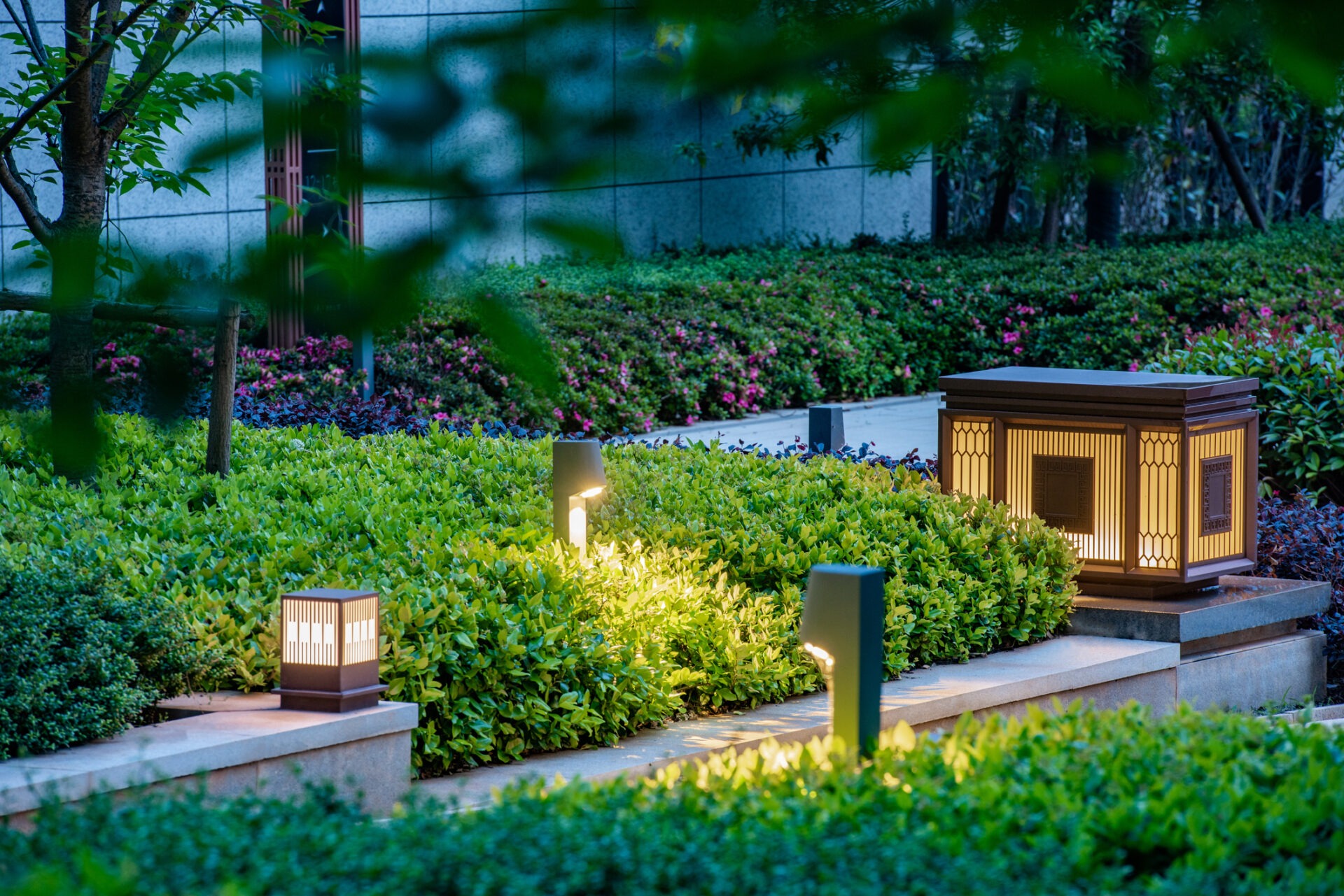 Illuminated garden path with glowing lanterns and lush greenery, surrounded by a modern stone structure, creating a serene, tranquil atmosphere.