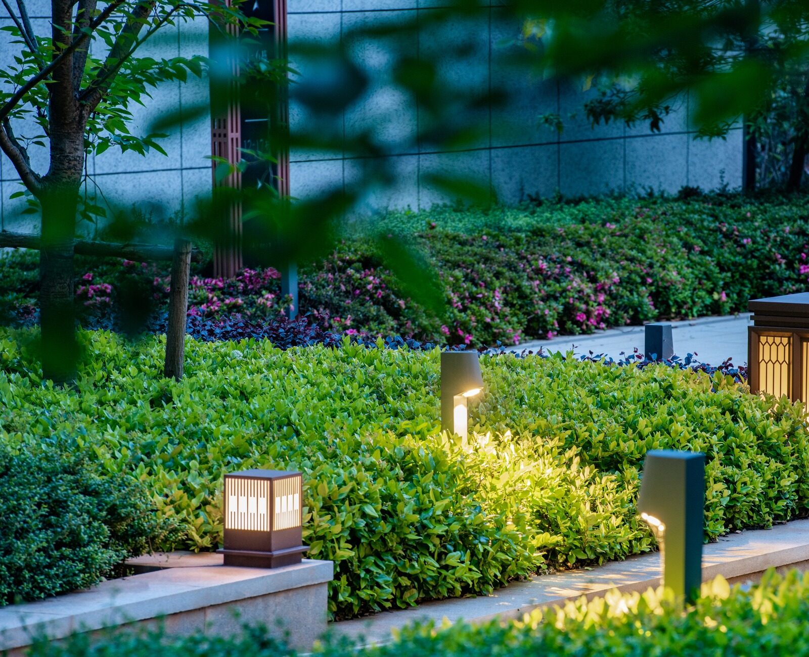 Illuminated garden path with glowing lanterns and lush greenery, surrounded by a modern stone structure, creating a serene, tranquil atmosphere.