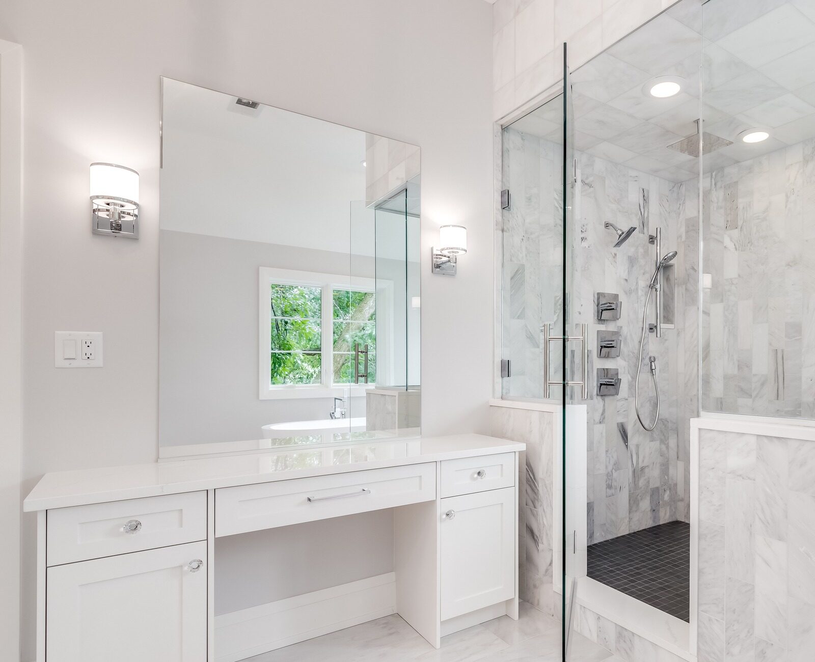 Modern bathroom with white marble tiles, glass shower enclosure, and a vanity mirror. Bright lighting and window view add natural ambiance.