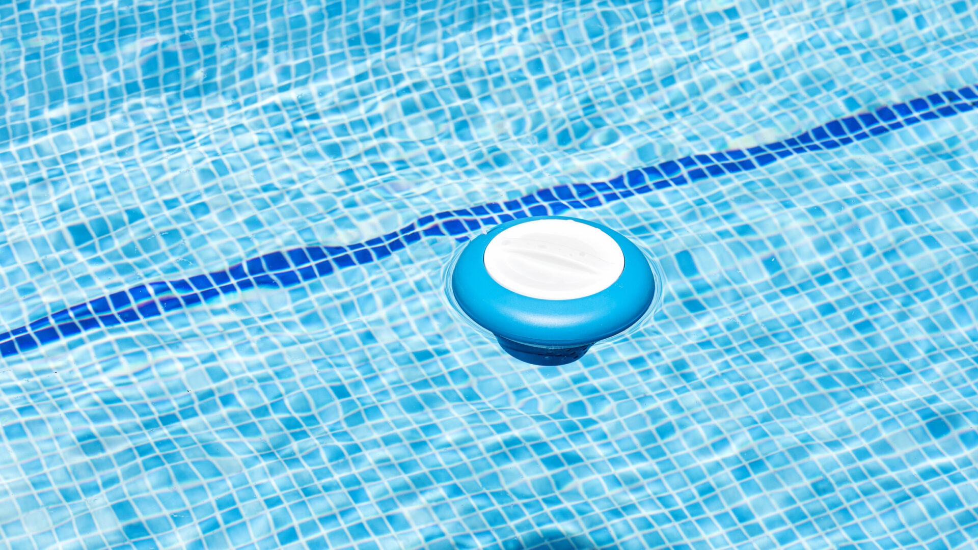 A blue pool chlorinator floats on clear water tiles, casting a shadow under bright sunlight. The scene is peaceful and summery.