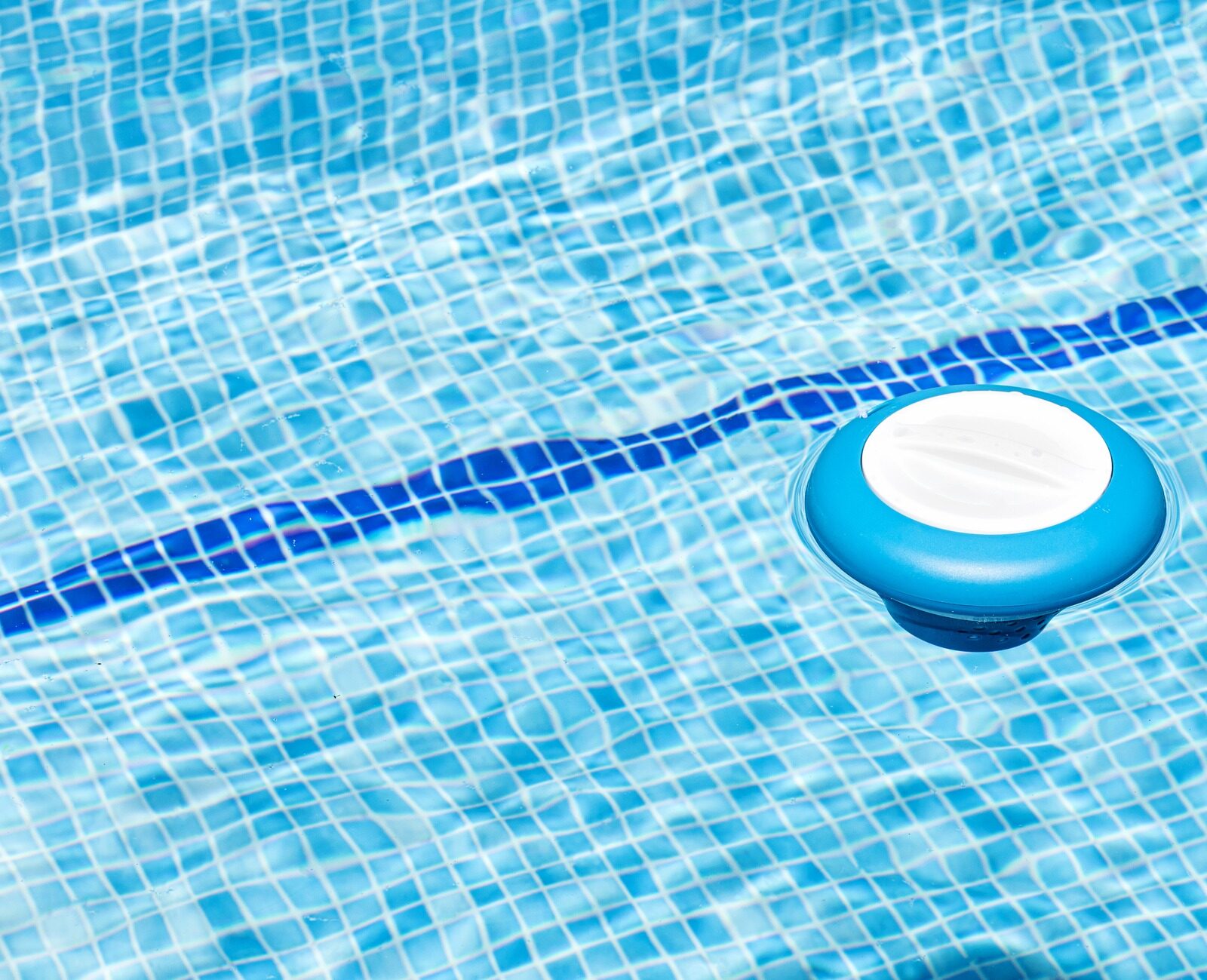 A blue pool chlorinator floats on clear water tiles, casting a shadow under bright sunlight. The scene is peaceful and summery.