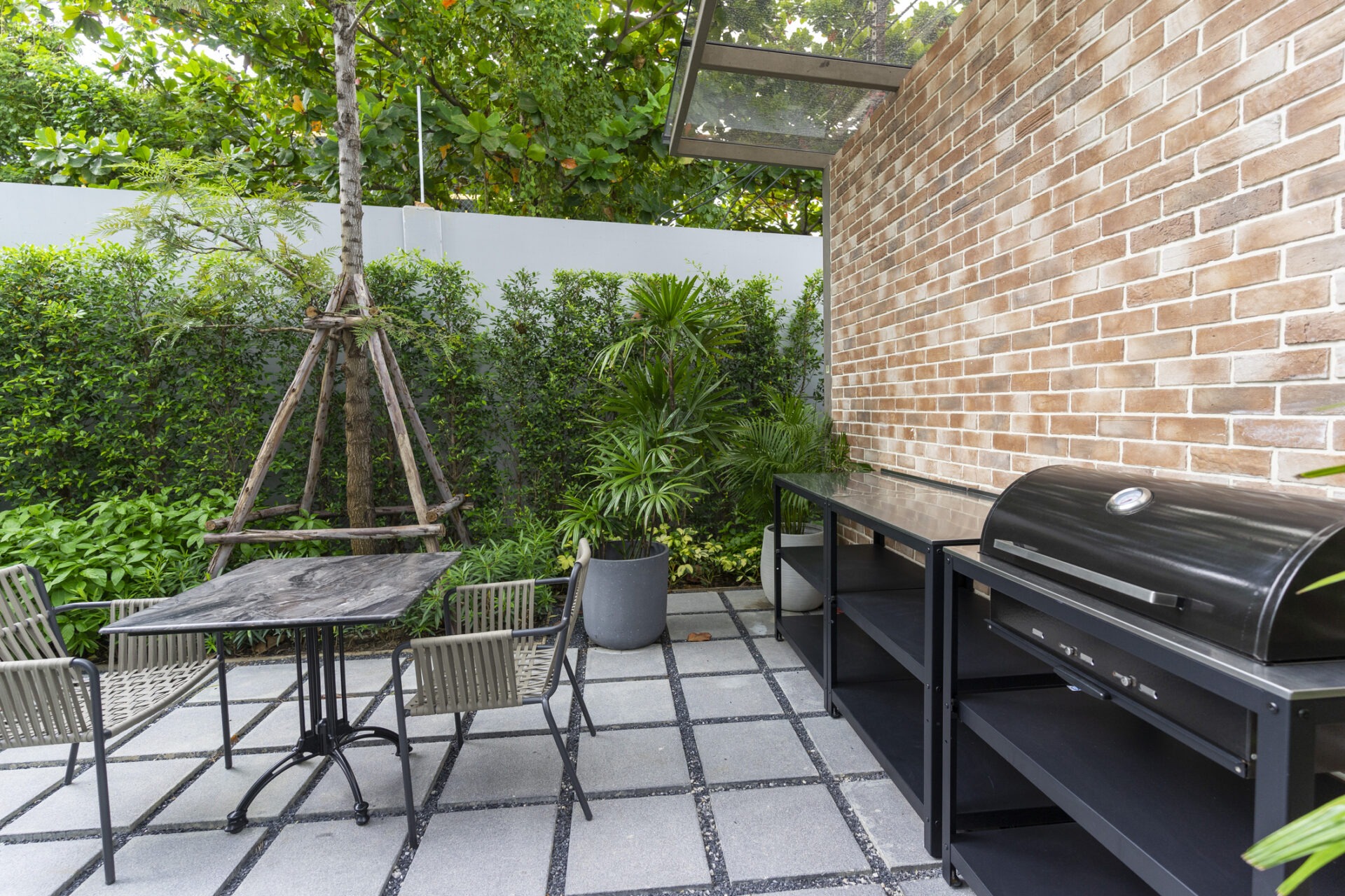 Outdoor patio with grill, brick wall, and marble table. Surrounded by lush greenery and modern metal furniture. Peaceful garden setting.