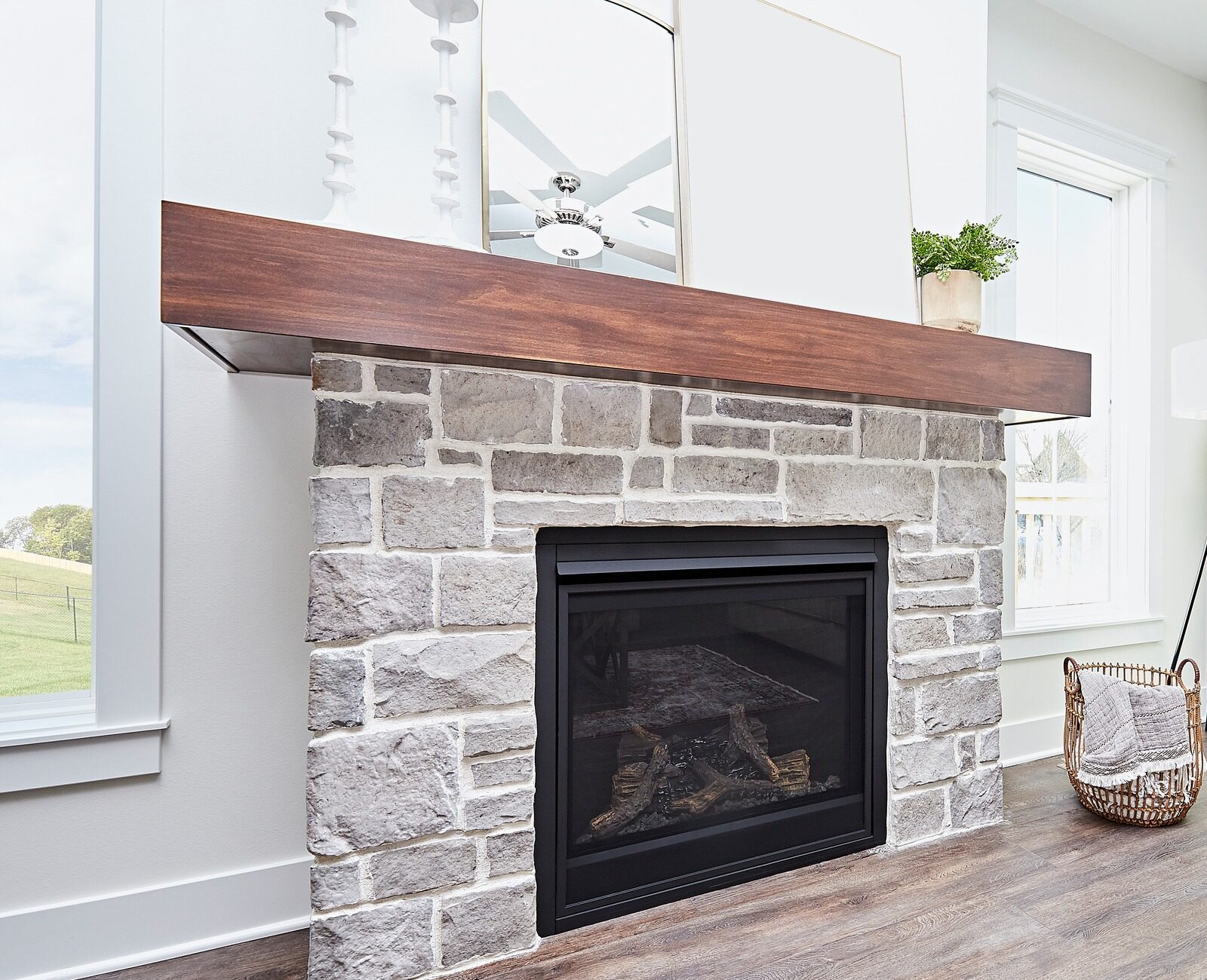 A cozy living room features a stone fireplace, wooden mantel, basket, floor lamp, and beige couch with yellow pillow. Sunlight filters through windows.