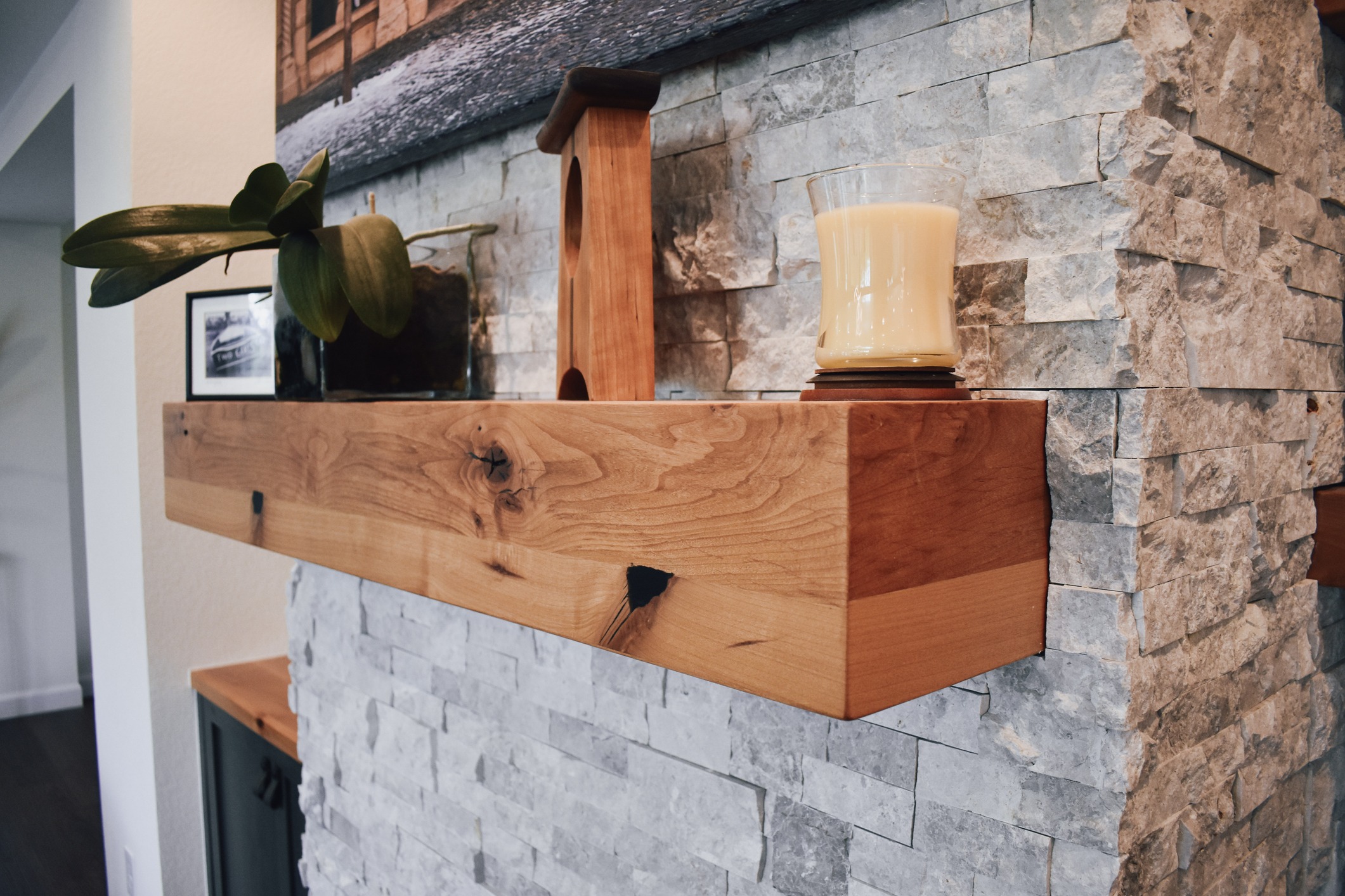 A wooden mantel decorates a stone fireplace with a candle, framed photo, and a potted plant enhancing the cozy, rustic interior.