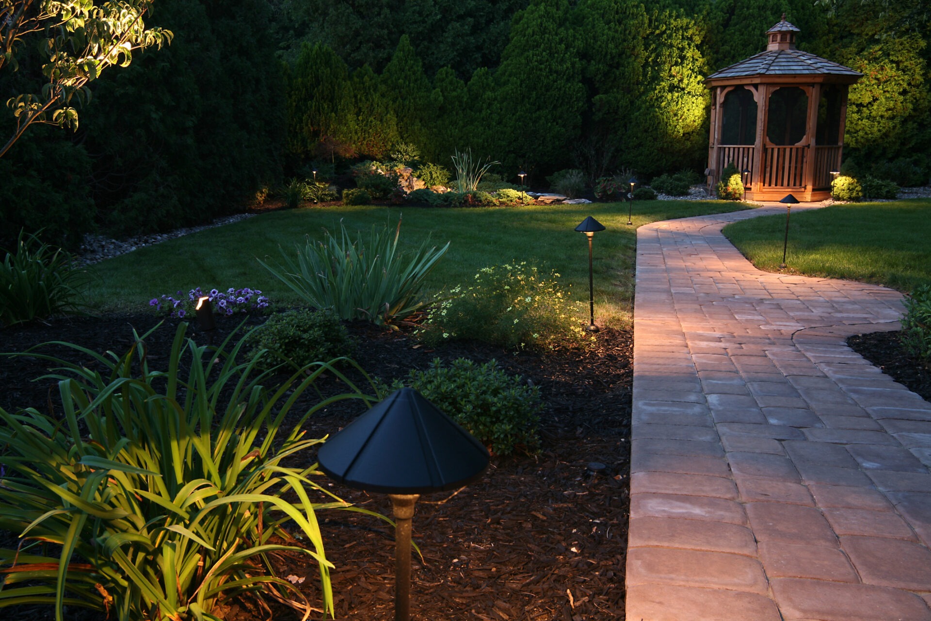 A serene garden with lush greenery, illuminated walkway lights, and a charming wooden gazebo in the background, creating a peaceful evening atmosphere.