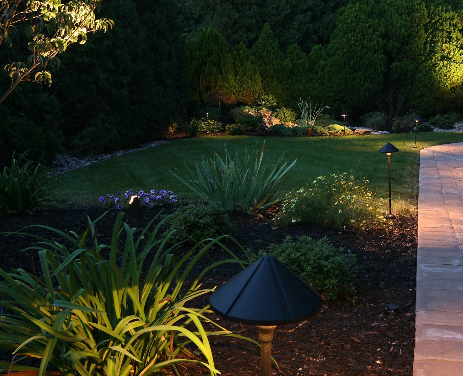 A serene garden with lush greenery, illuminated walkway lights, and a charming wooden gazebo in the background, creating a peaceful evening atmosphere.