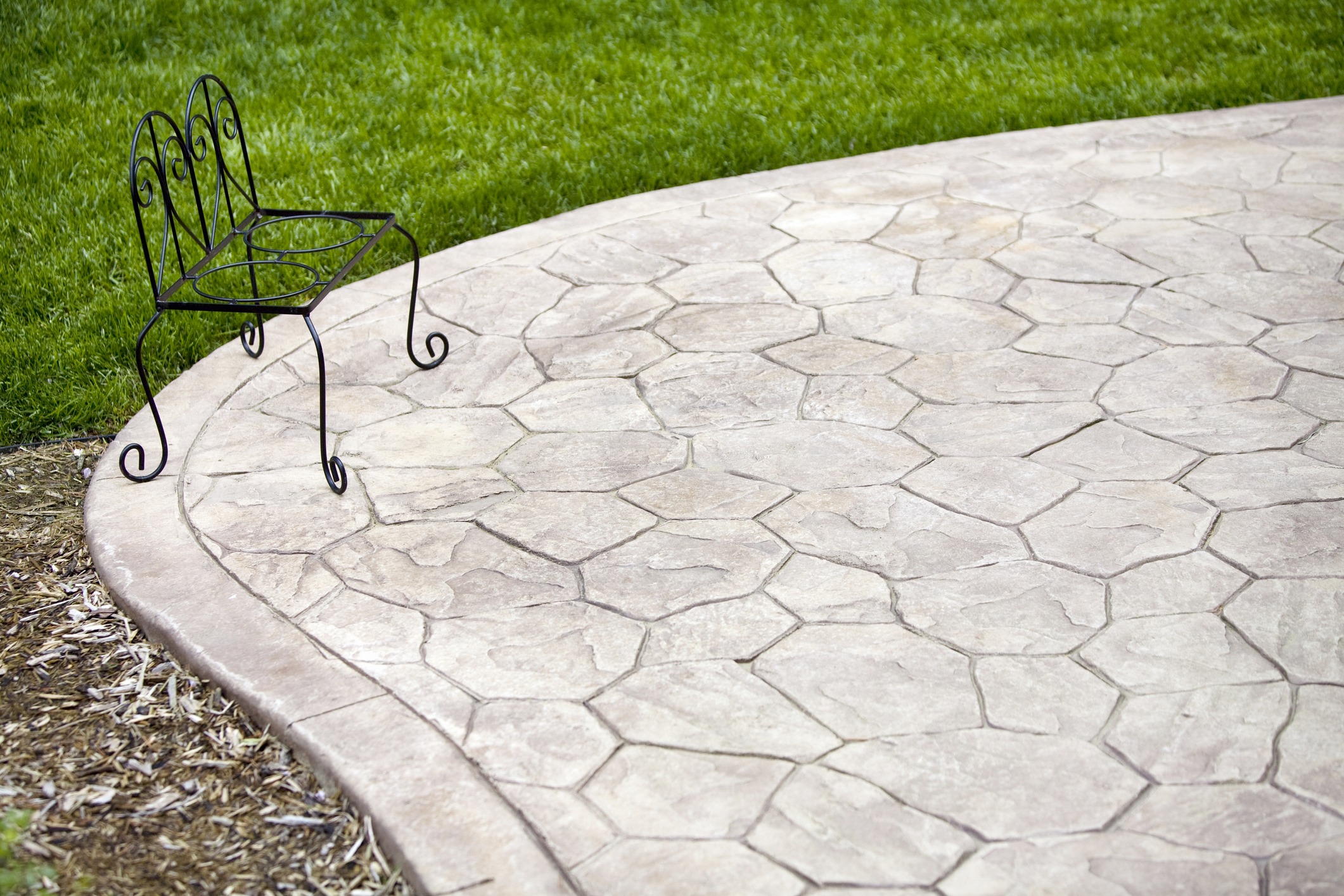 A decorative metal bench sits on a patterned stone patio, surrounded by neatly trimmed green grass and mulch, in an outdoor setting.