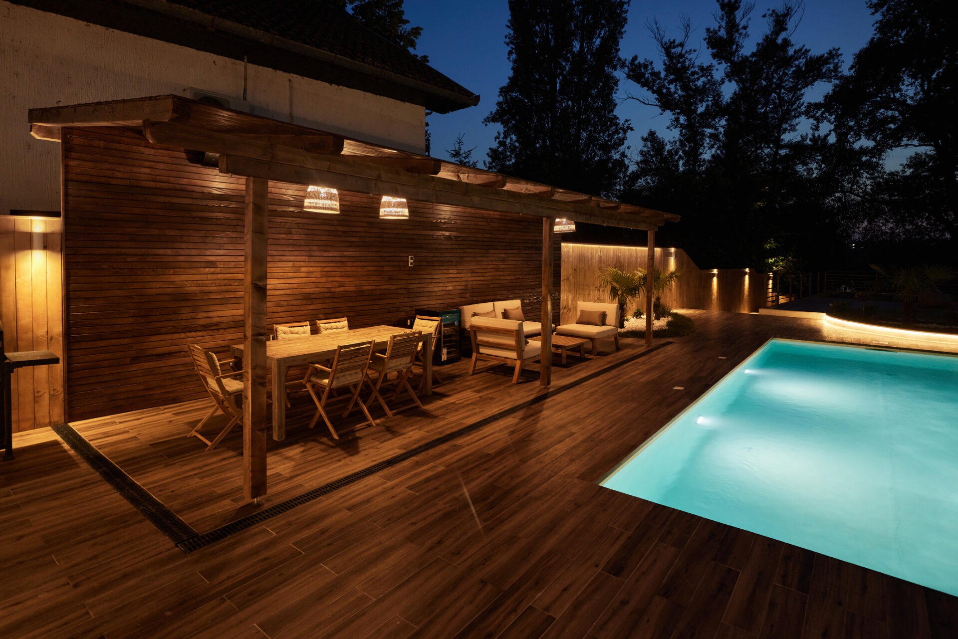 A well-lit outdoor patio features wooden furniture, a modern pergola, and a pool, surrounded by trees under a dusky evening sky.