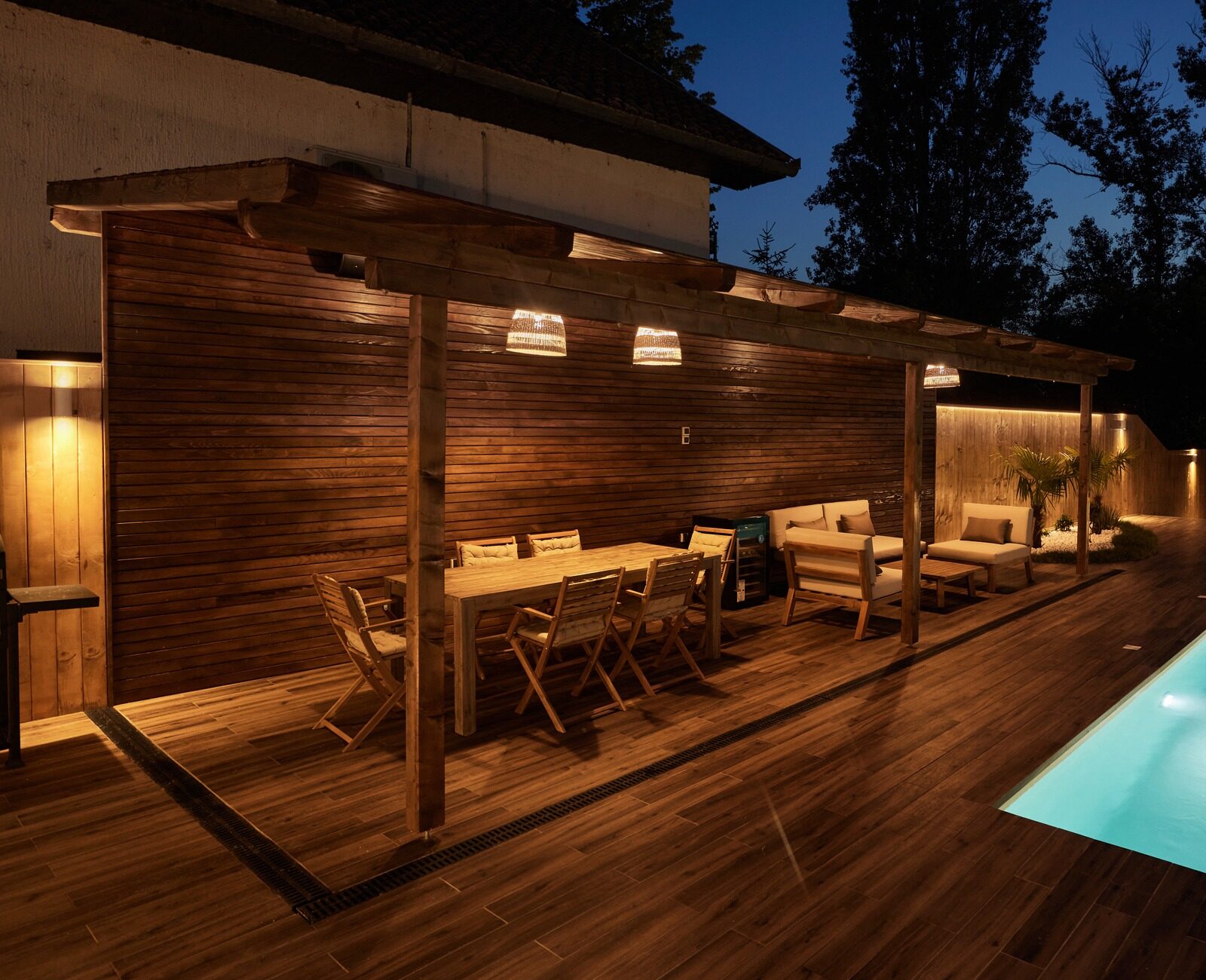 A well-lit outdoor patio features wooden furniture, a modern pergola, and a pool, surrounded by trees under a dusky evening sky.