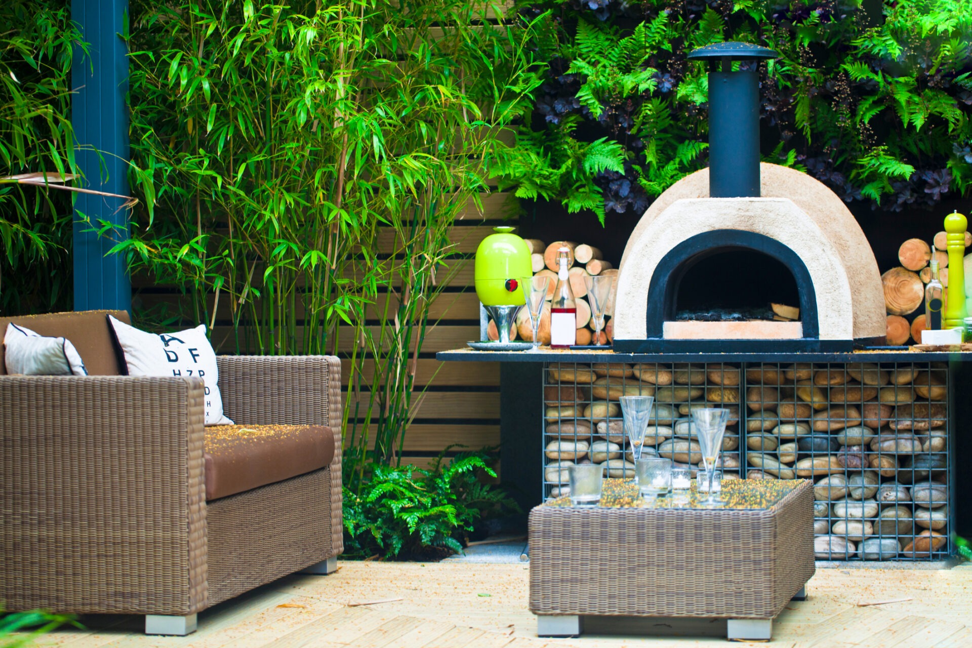Outdoor patio features wicker furniture, a stone pizza oven, and lush green bamboo, creating a cozy, inviting garden atmosphere for relaxation.