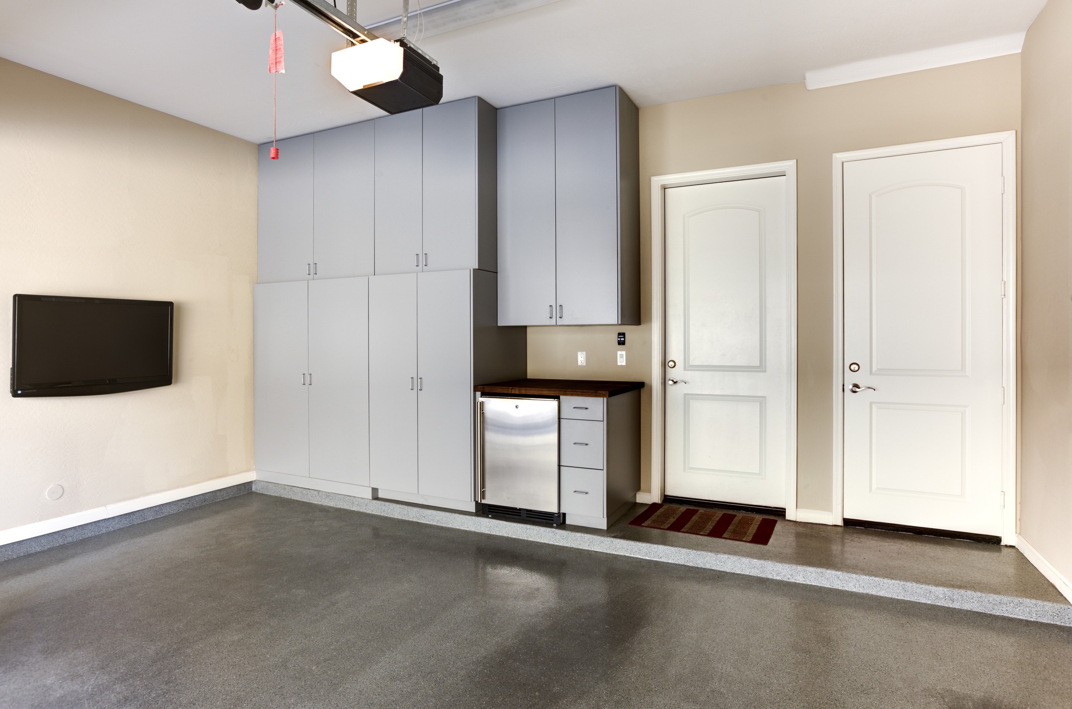 Organized garage with gray cabinets, wall-mounted TV, and stainless steel mini fridge. Two closed white doors lead to other rooms. Clean, polished floor.