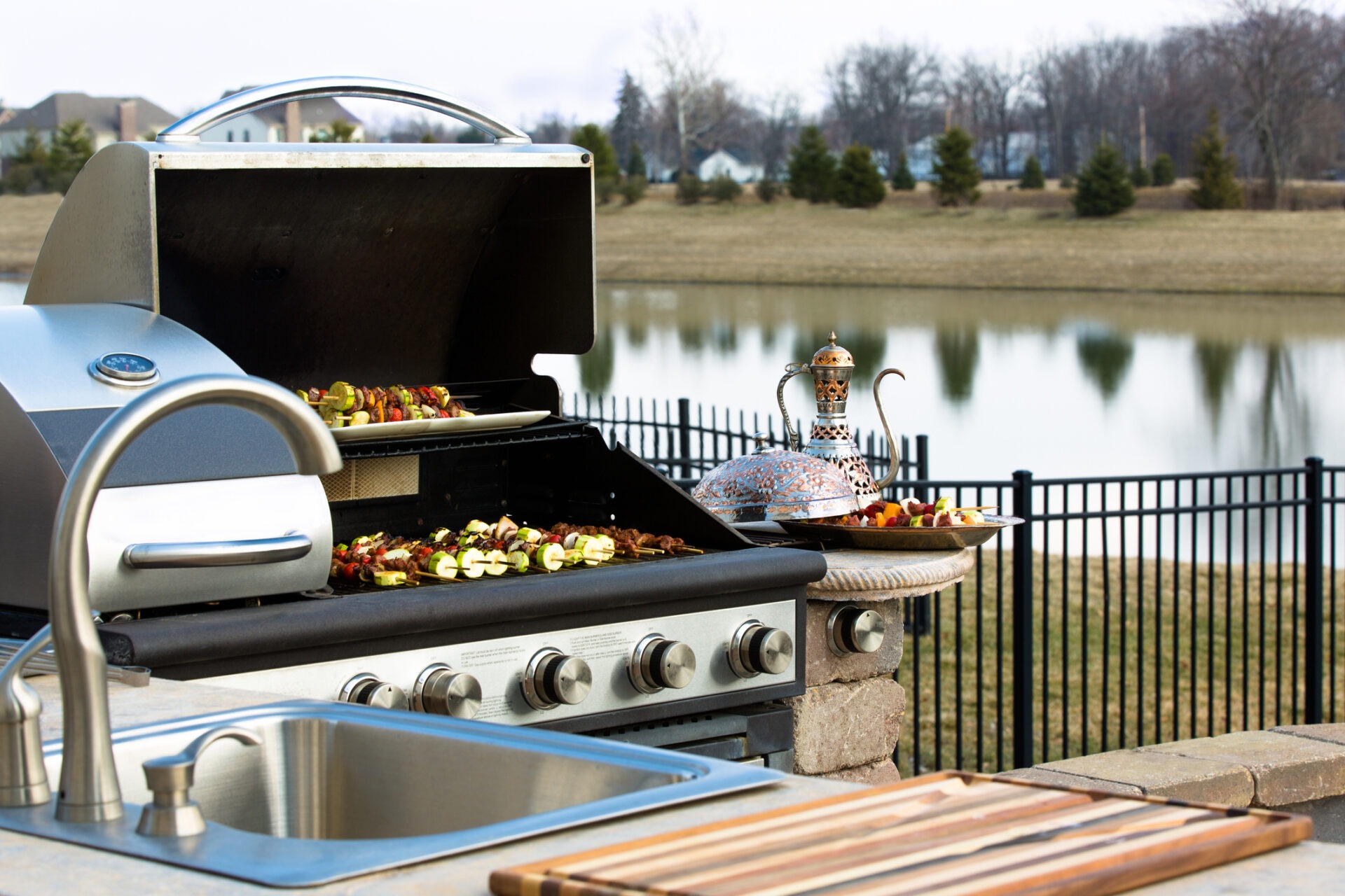 A barbecue and sink setup near a pond with skewered vegetables grilling, decorative pot, and a platter beside a metal fence.