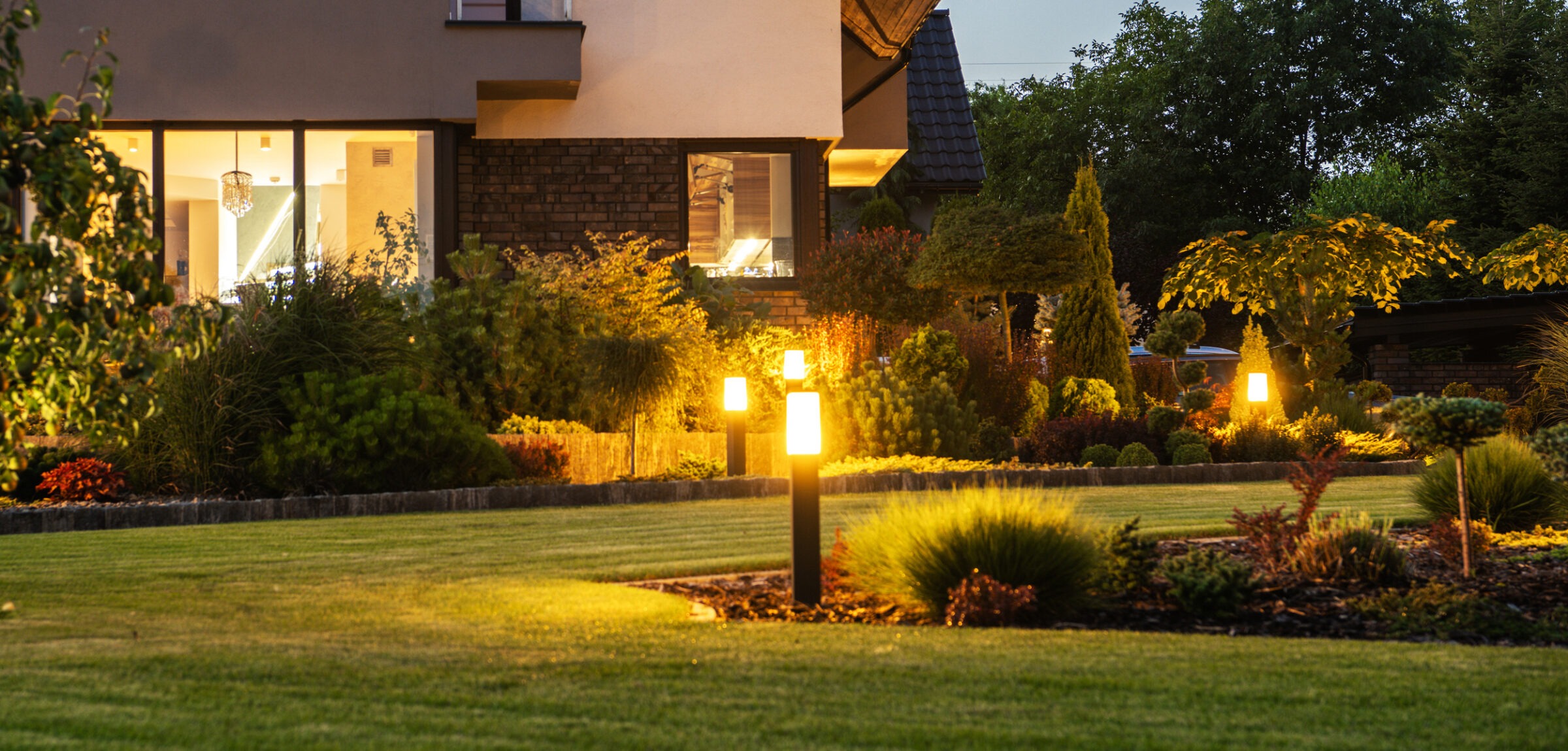Modern house with illuminated garden at dusk, featuring well-manicured lawn, neatly arranged shrubs, and warm lighting creating a serene evening ambiance.