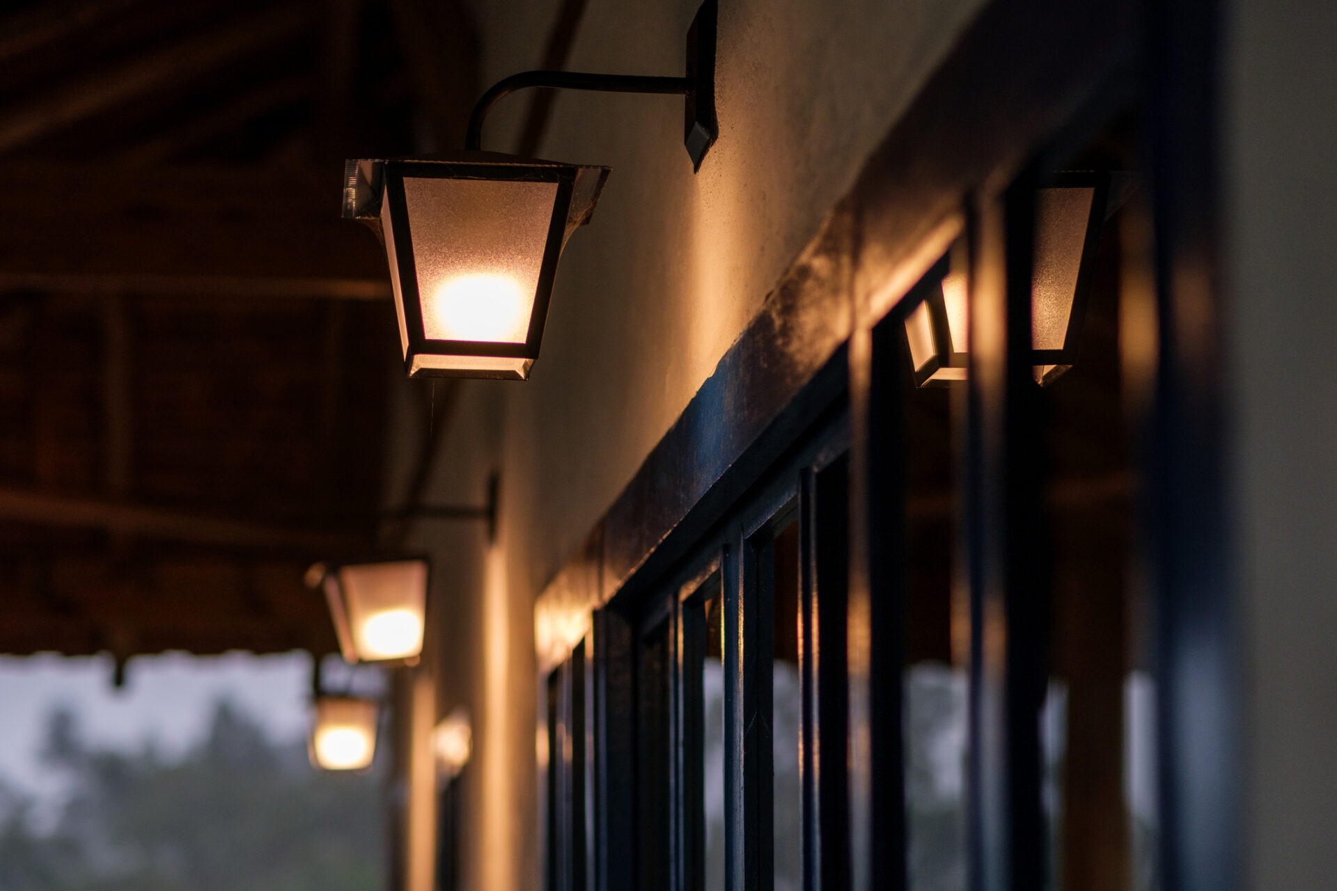 Warmly lit exterior lamps line a building's wooden overhang, casting soft light on a dark glass window with blurred greenery in the background.